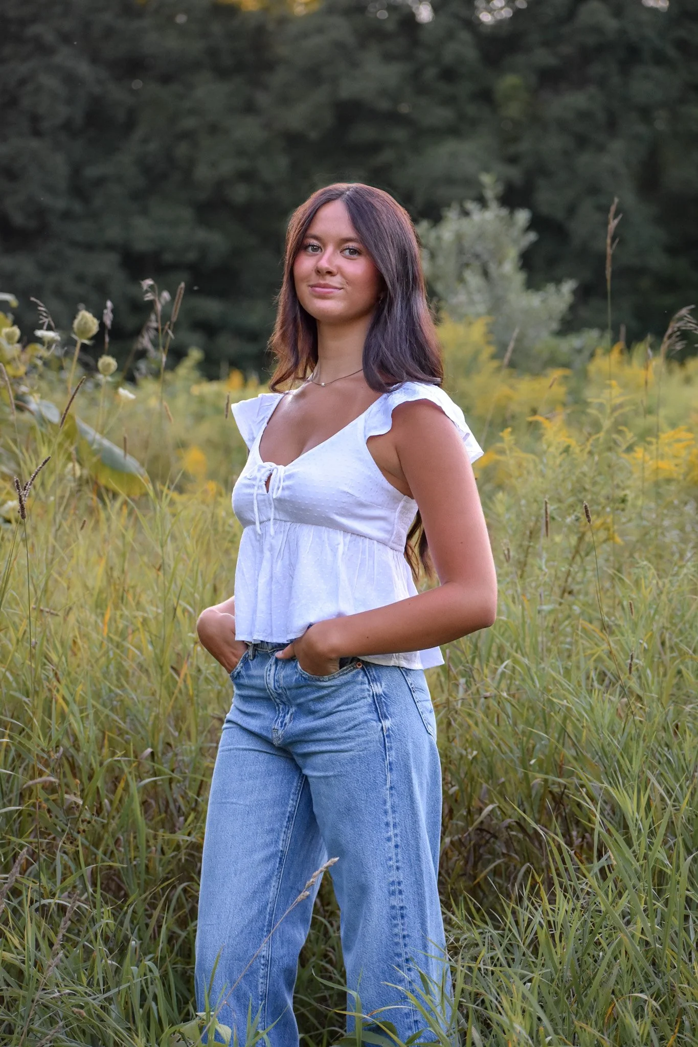 A young woman with long brown hair standing in a field of tall grass and wildflowers, dressed in a white top and blue jeans, with trees in the background.