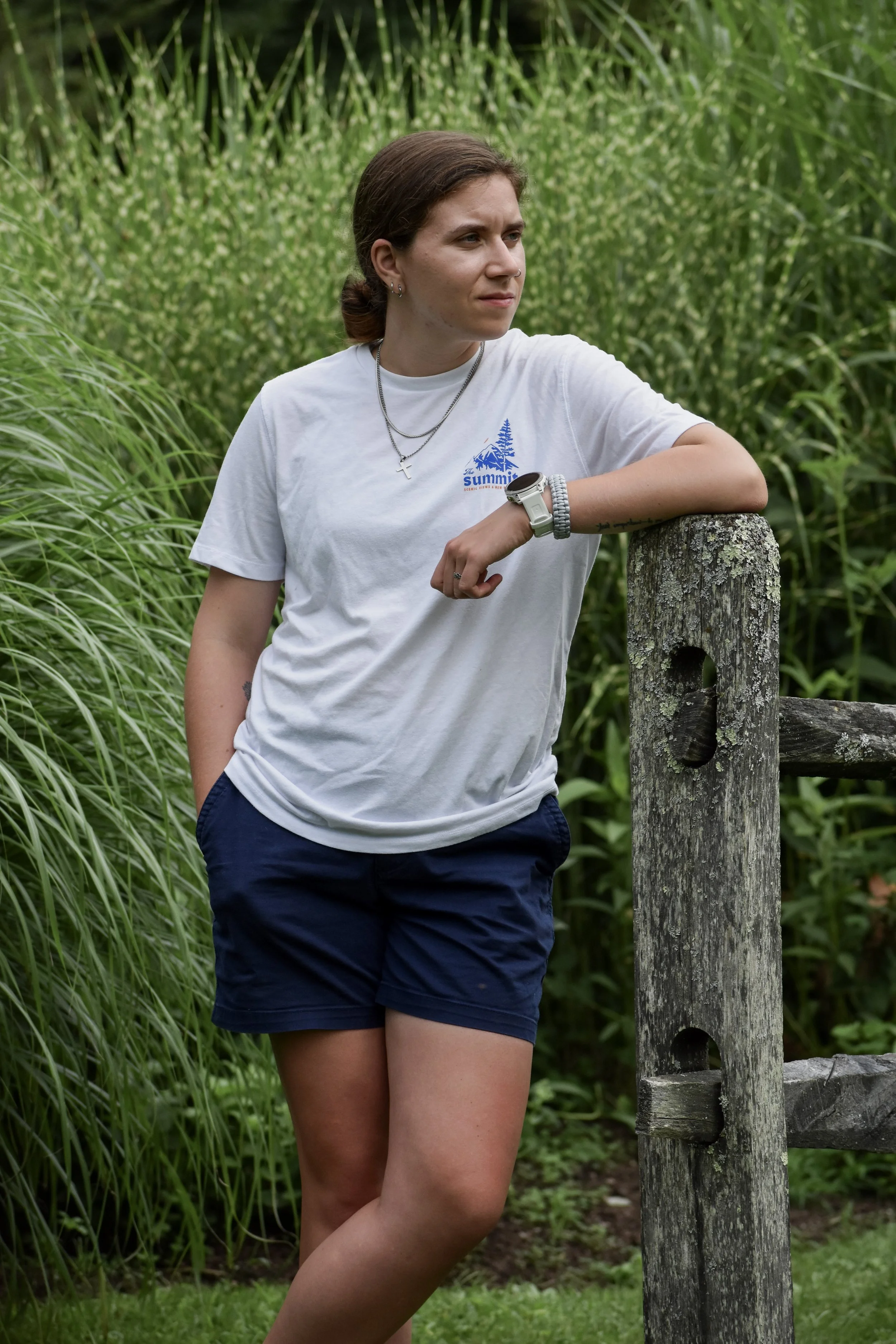 Young woman wearing a white t-shirt and navy shorts posing outdoors with one arm resting on a weathered wooden post, surrounded by tall green grass and foliage.