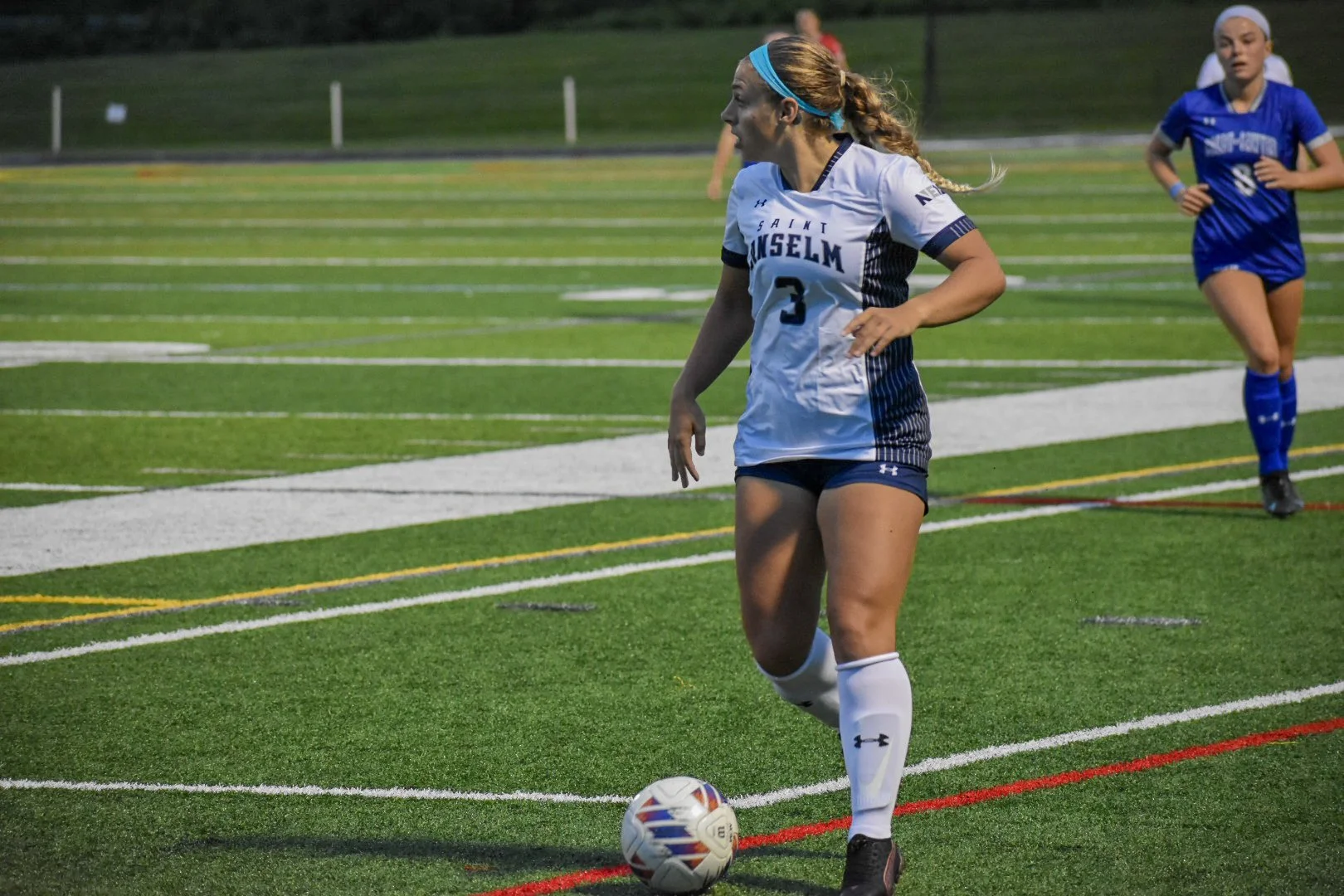 A female soccer player in a white and blue uniform with the number 3, standing on a green field with a soccer ball at her feet, preparing to kick or pass the ball.