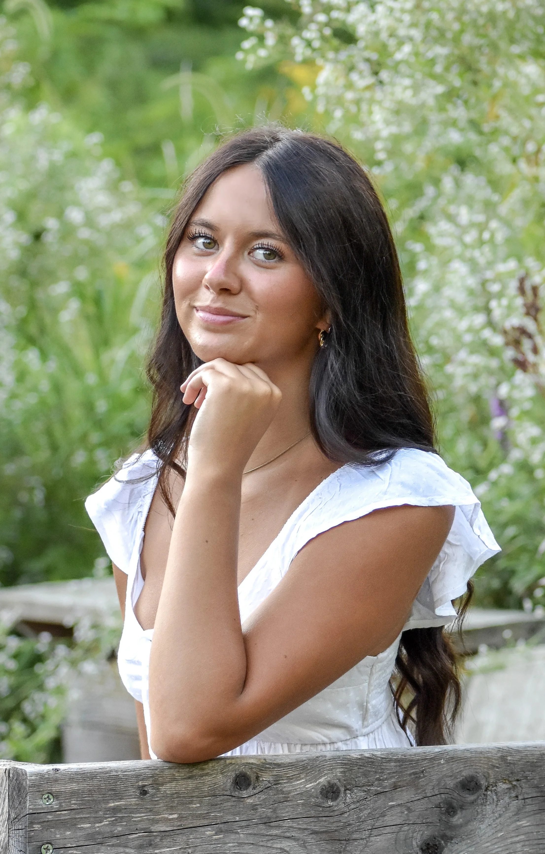 A young woman with long dark hair, wearing a white dress, posing outdoors with her chin resting on her hand and a gentle smile, background filled with green foliage and white flowers.