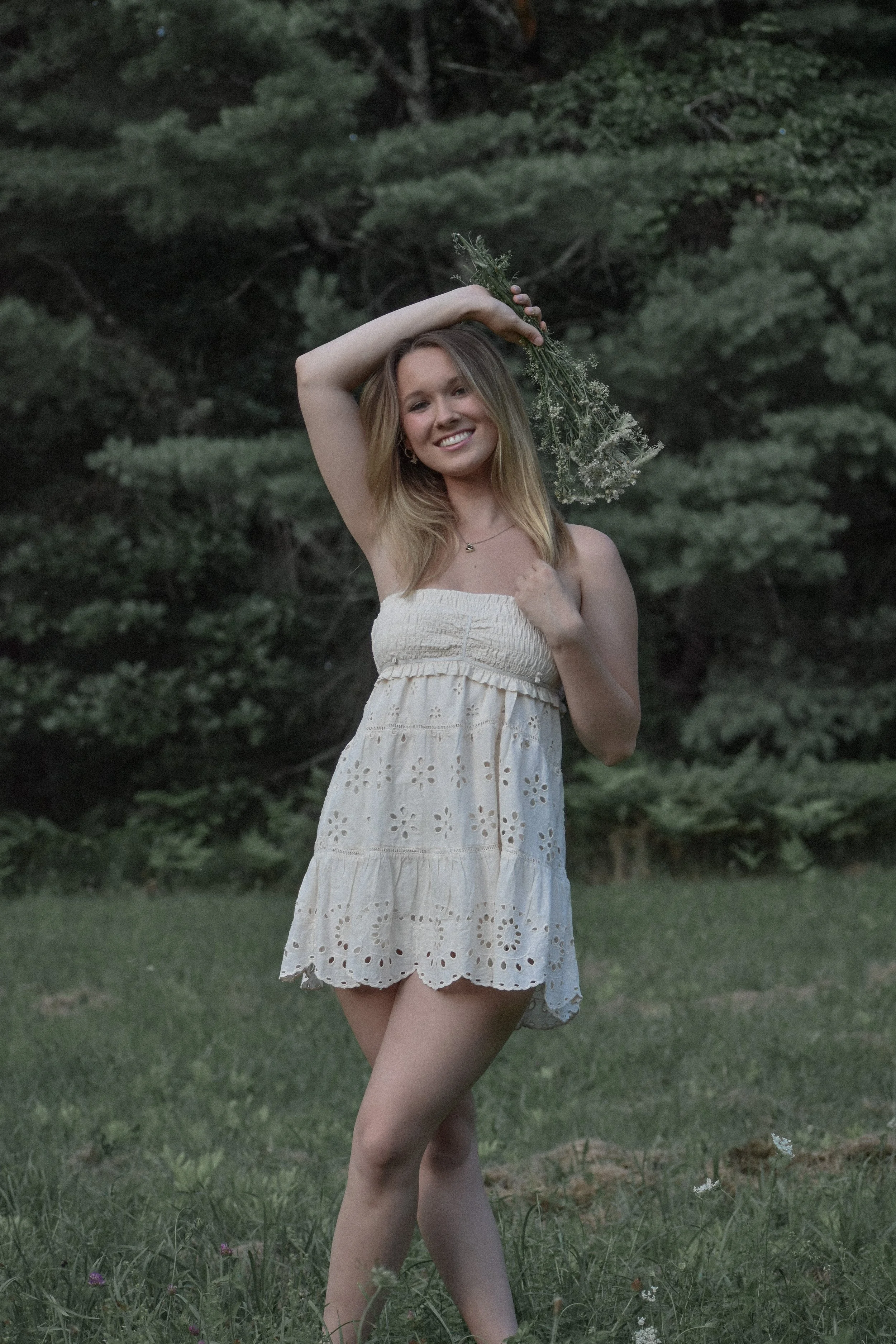 A young woman standing outdoors in a grassy area with trees in the background, holding a bunch of flowers over her shoulder, smiling, wearing a white strapless dress with eyelet details.