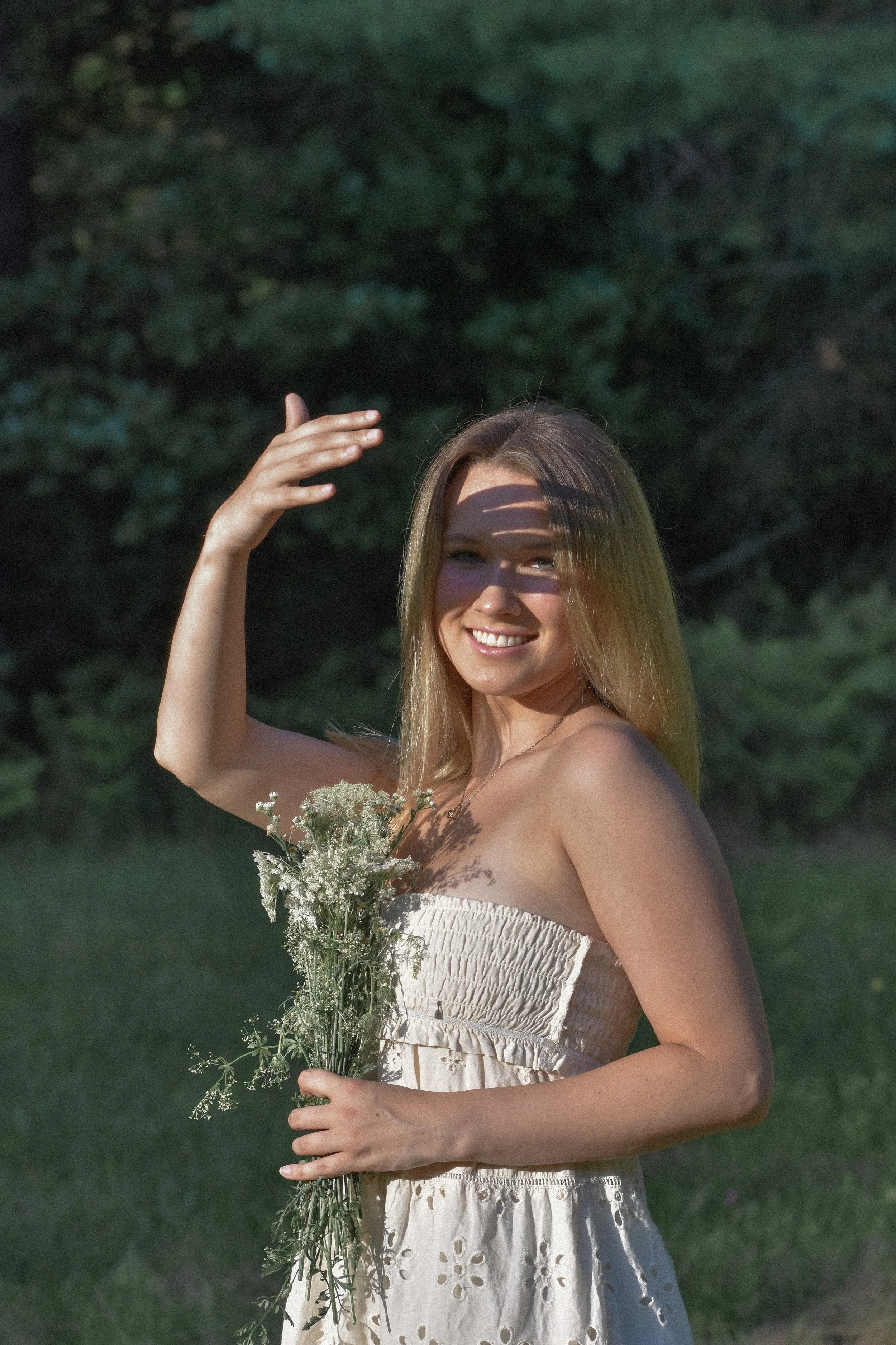 A young woman with long blonde hair smiling and holding a bouquet of white wildflowers outdoors in a green garden, with sunlight casting a shadow across her face.