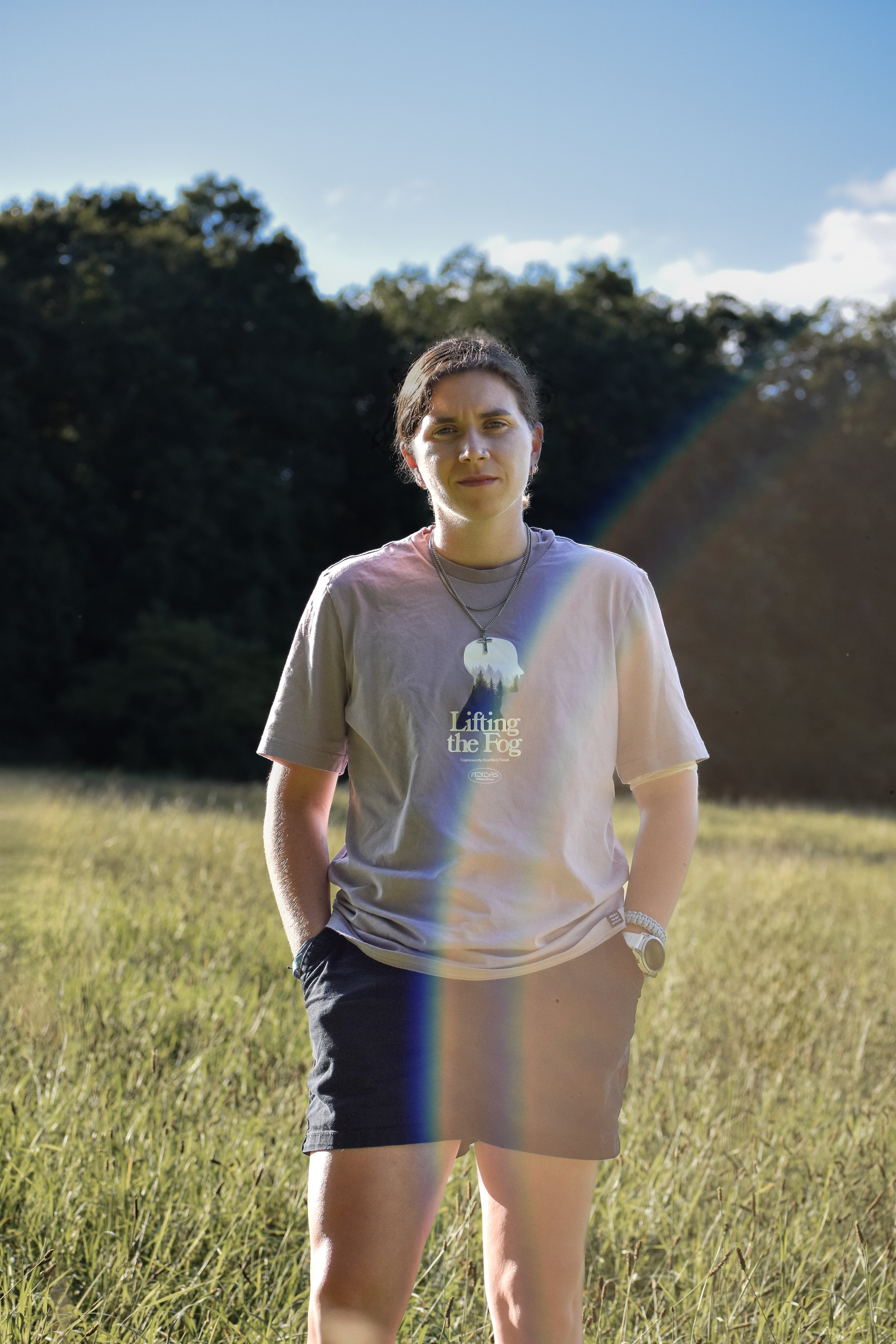 A woman standing in a grassy field with trees in the background and a rainbow-colored lens flare across her body, wearing a t-shirt with text and graphics, shorts, and jewelry.