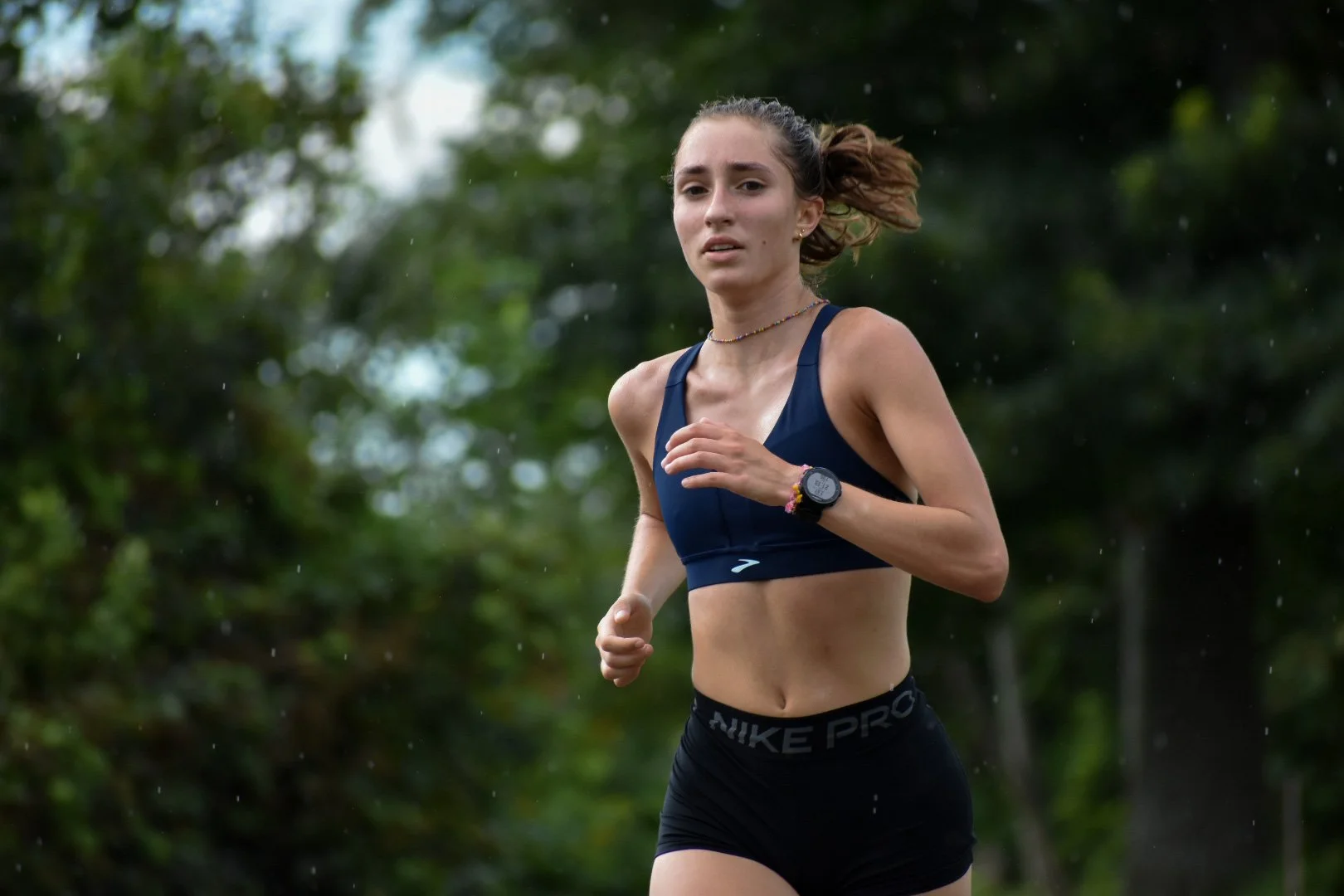 A young woman running outdoors in a race, wearing a navy sports bra, black Nike Pro shorts, and a sports watch, with trees in the background.