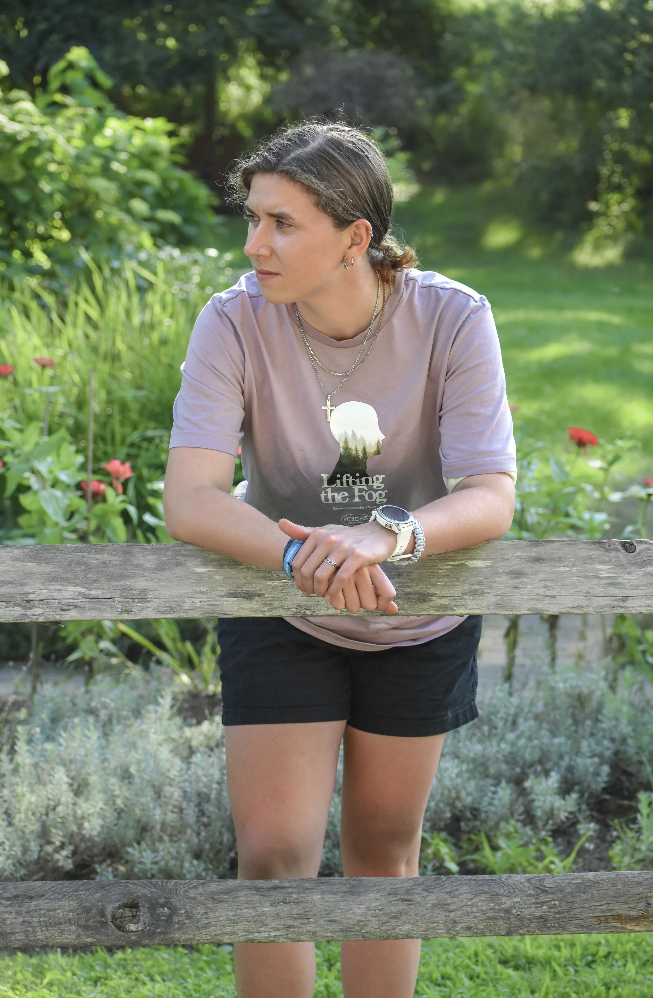 A young woman leaning on a wooden fence in a green outdoor setting, wearing a light purple t-shirt with a silhouette of a person's face and the text 'Lifting the Fog', black shorts, and jewelry, with trees and plants in the background.