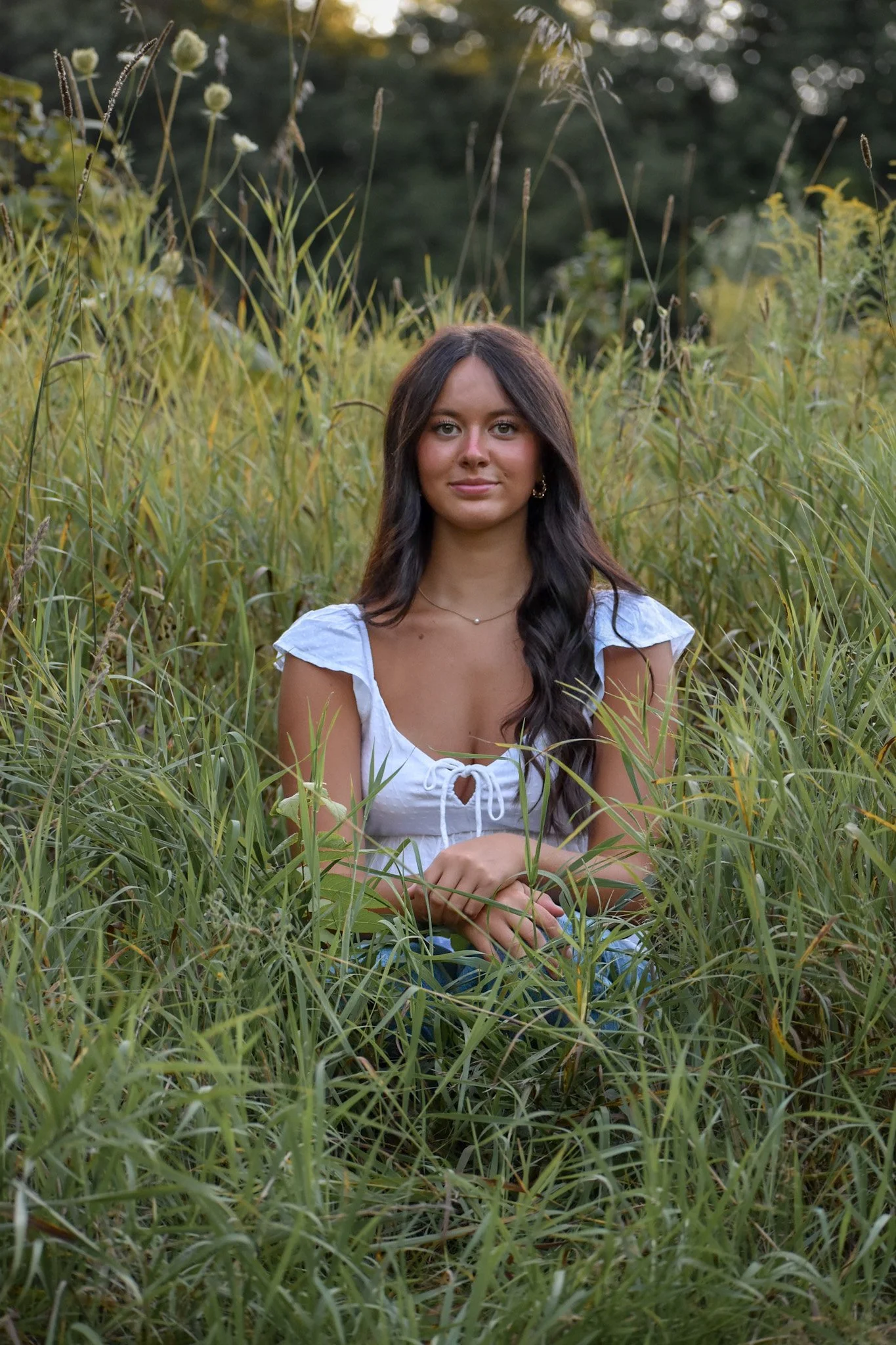A young woman with long dark hair, wearing a white top, sits in tall green grass outdoors with a blurred background of trees and plants, during sunset.
