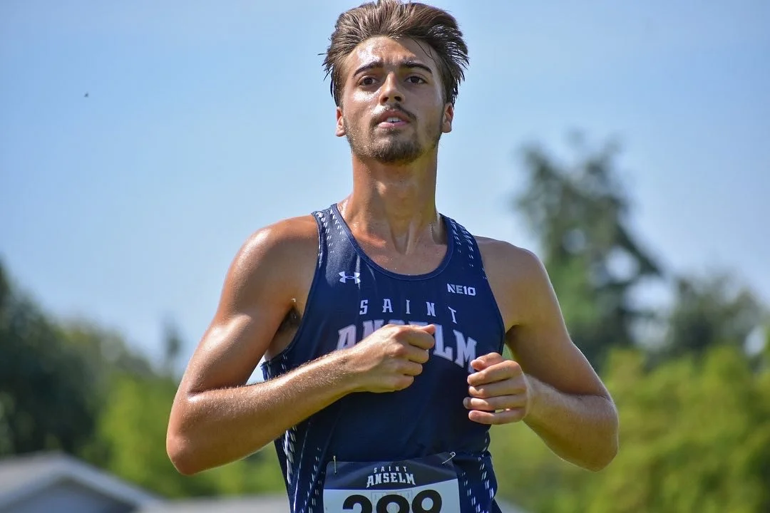 Young male athlete running outdoors, wearing a blue Saint Anselm sports jersey, with a race number 288, on a sunny day.