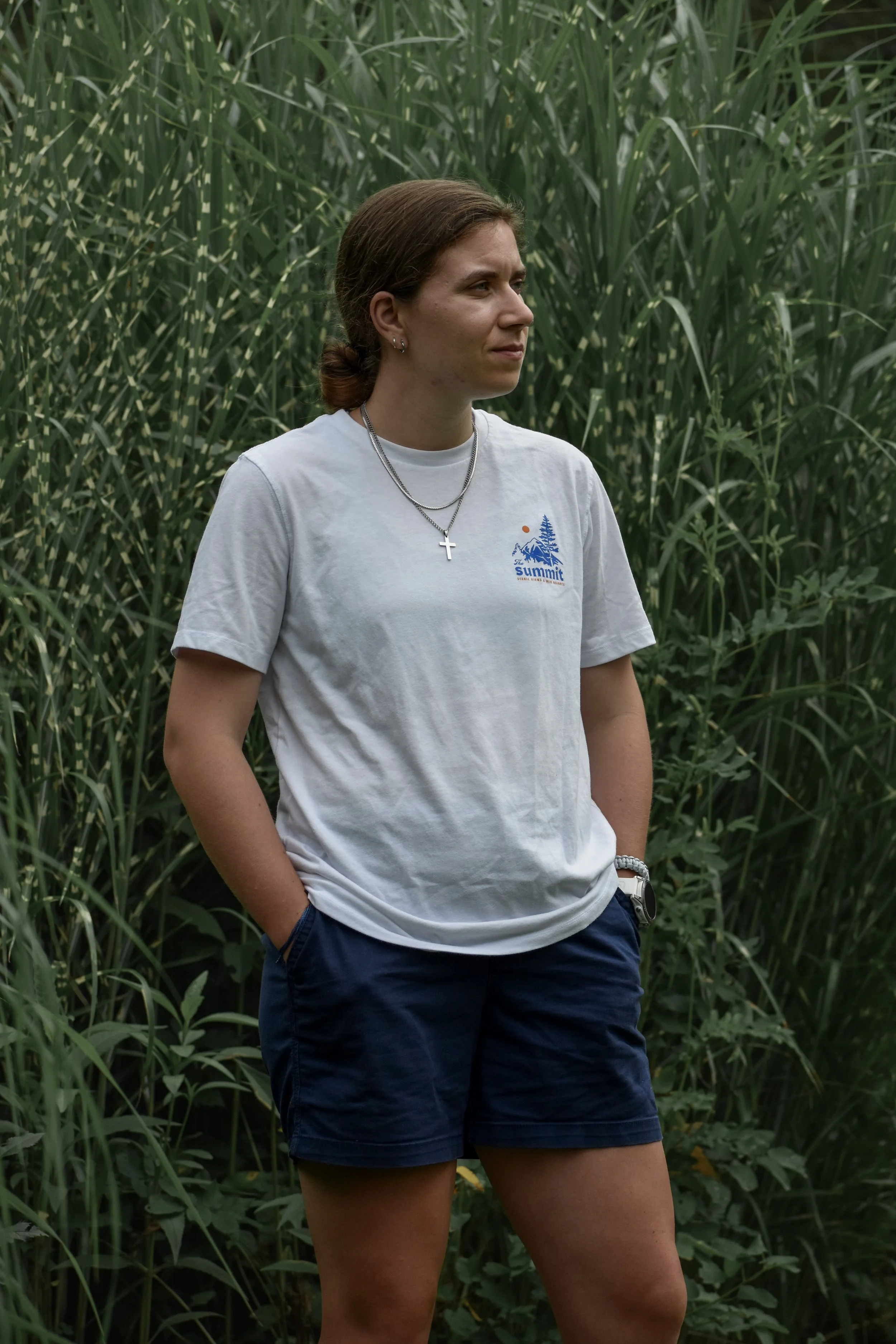 A woman with brown hair tied back, wearing a white t-shirt with a logo and navy blue shorts, standing among tall green grass.