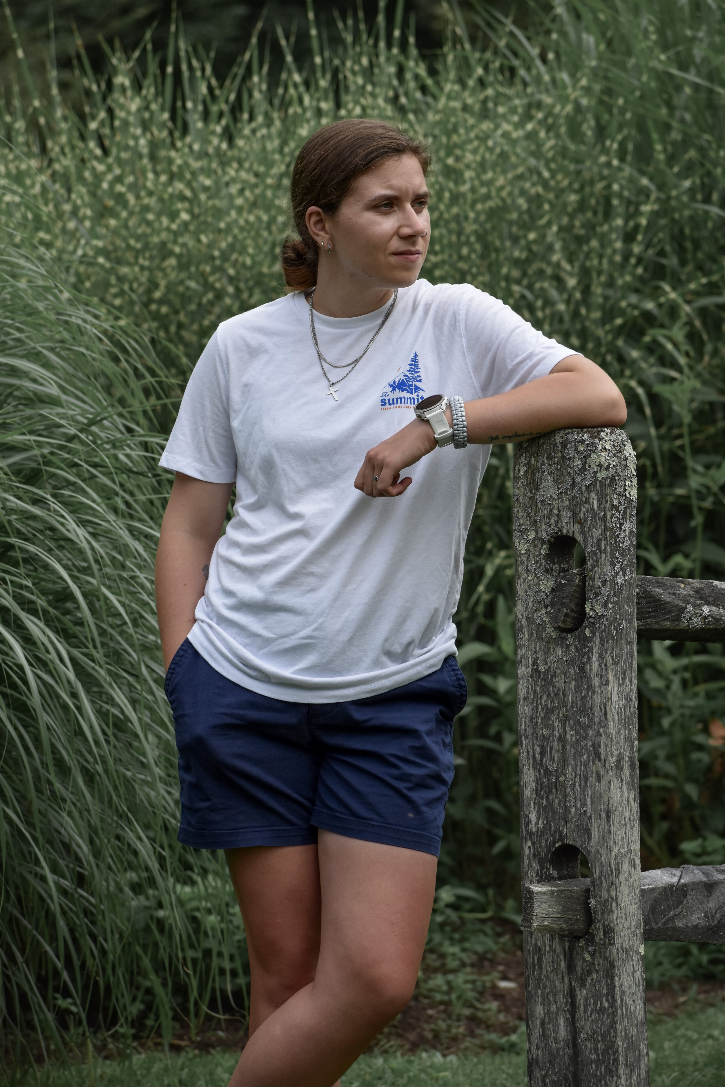Woman leaning against a weathered wooden post outdoors, surrounded by tall green grass and foliage.