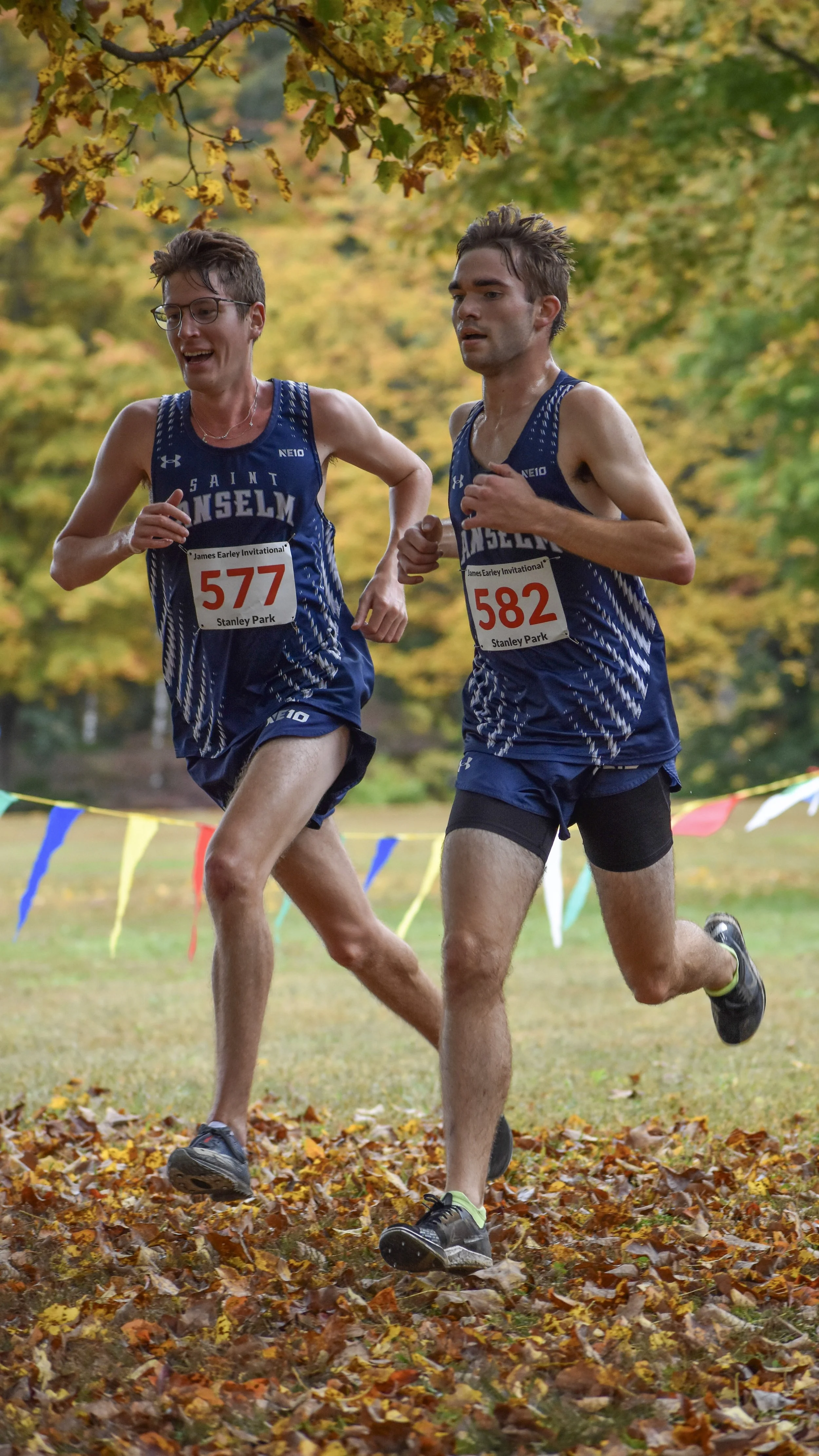 Two male runners participating in a cross-country race, wearing blue athletic uniforms with the word 'SAINT' visible, numbered bibs 577 and 582, running on a leaf-covered trail with autumn trees in the background.