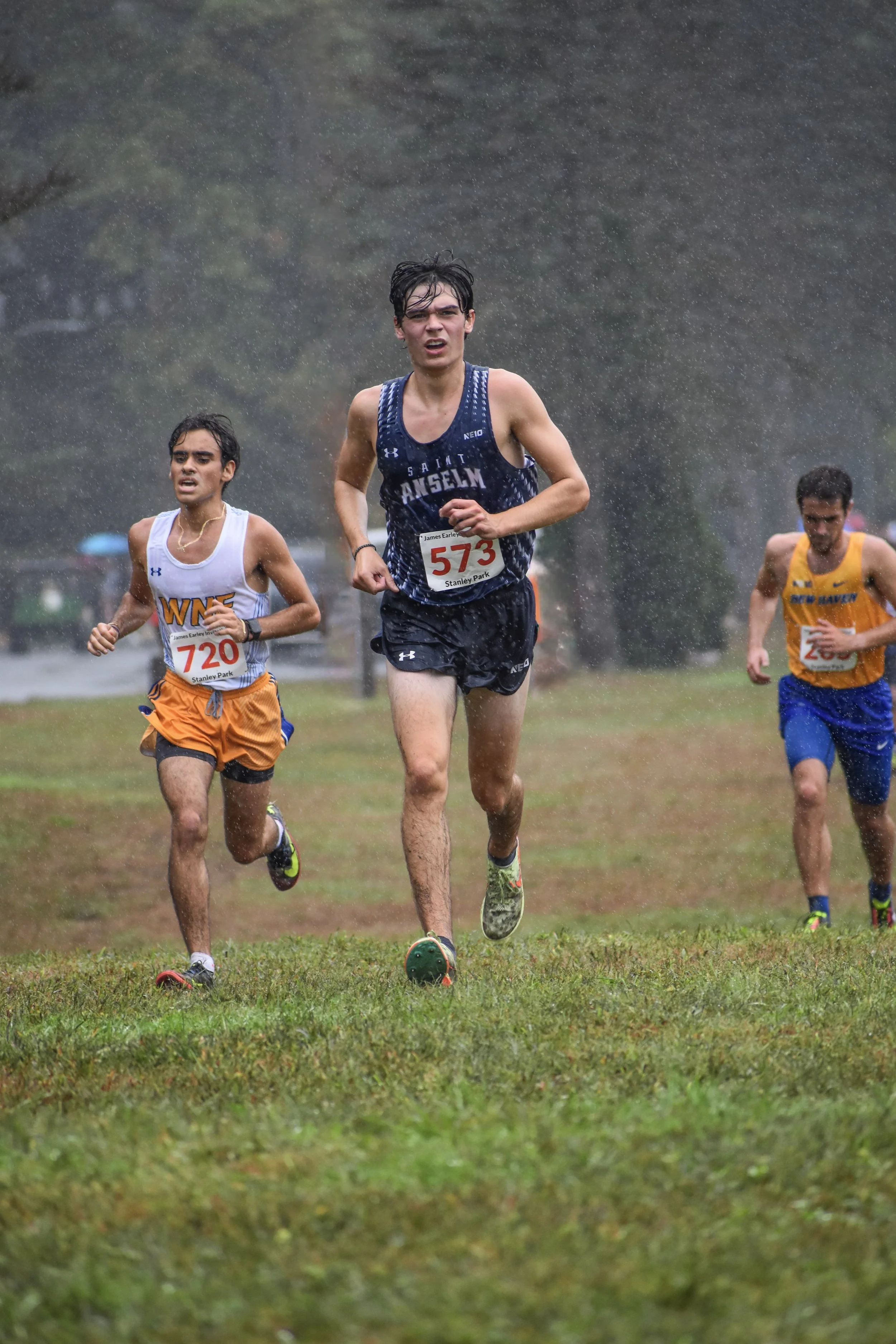 Young male runners competing in a cross-country race in the rain on a grassy field, with trees in the background.