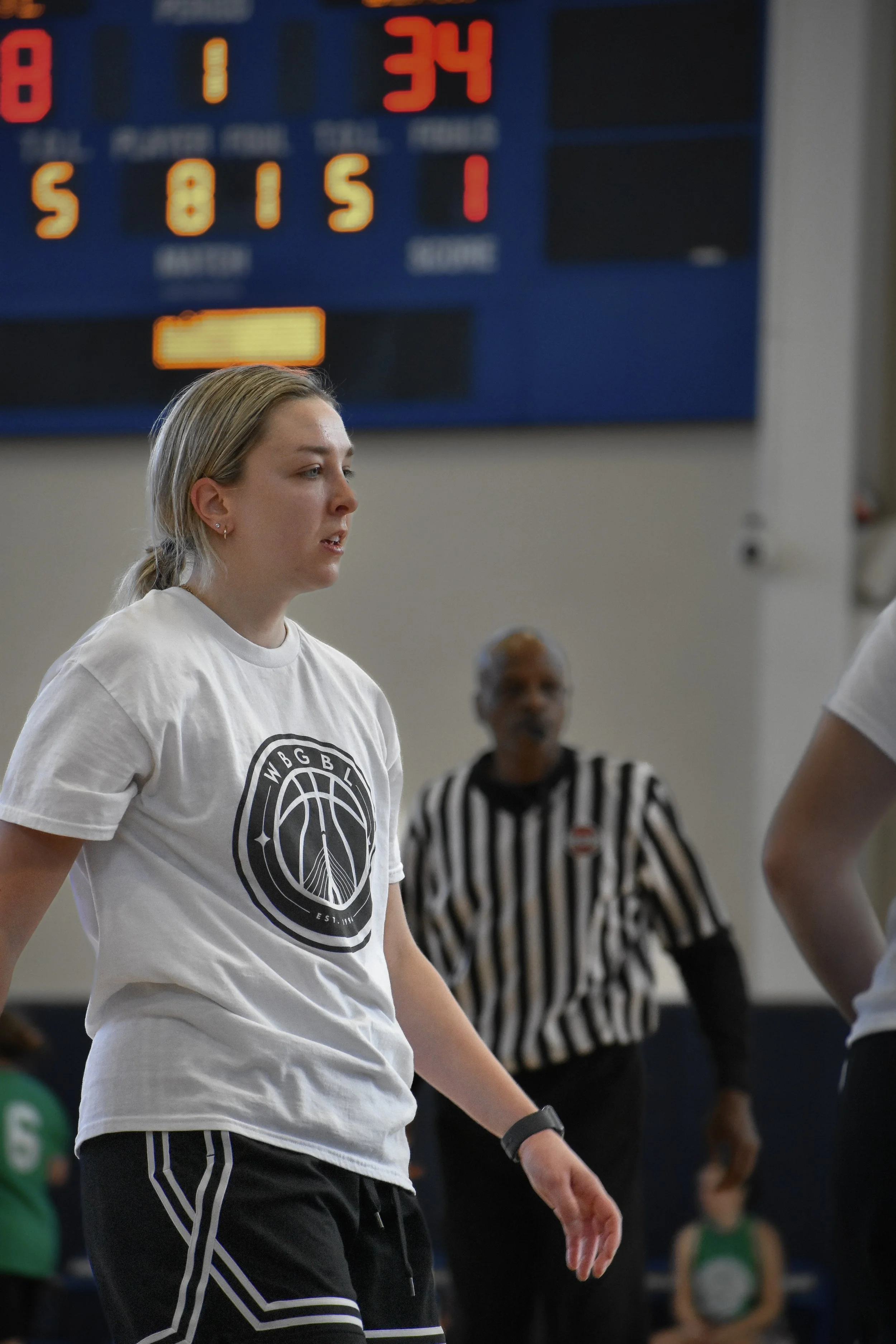 A young female basketball player stands on the court during a game, wearing a white t-shirt with a Brooklyn Nets logo and black athletic shorts. In the background, a referee in a black and white striped shirt and a digital scoreboard are visible.