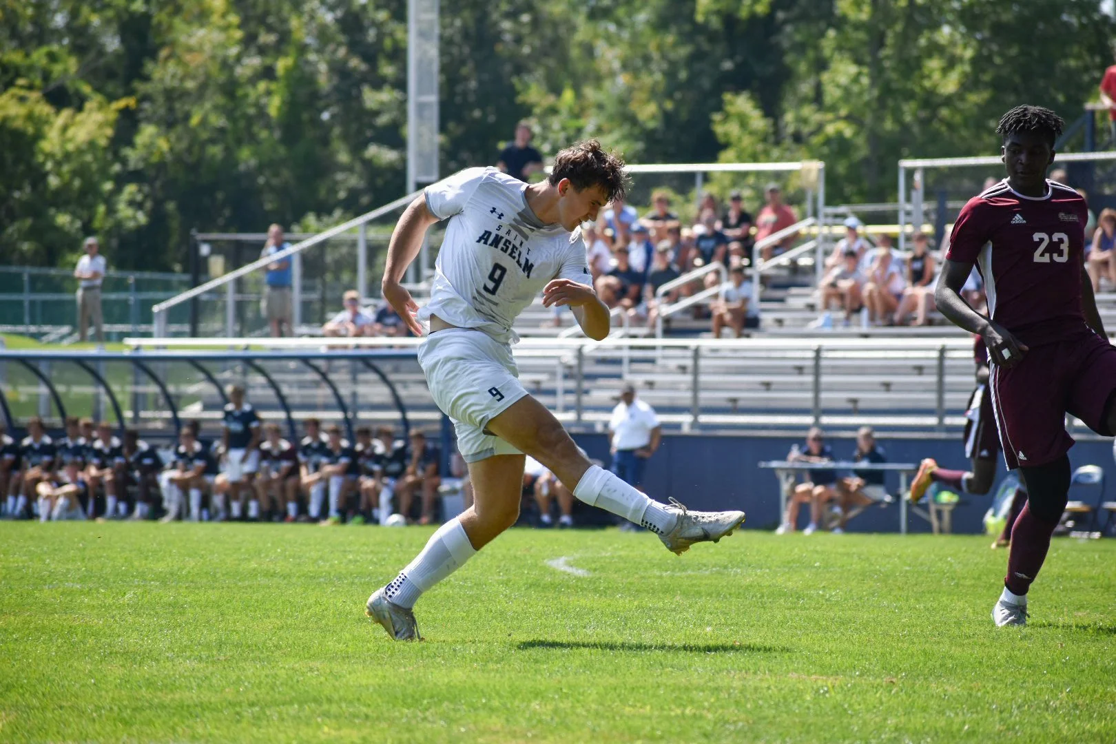 A soccer player in a white uniform with the number 9 is kicking a soccer ball on a green field during a match, with spectators in the bleachers and a player in a maroon uniform nearby.