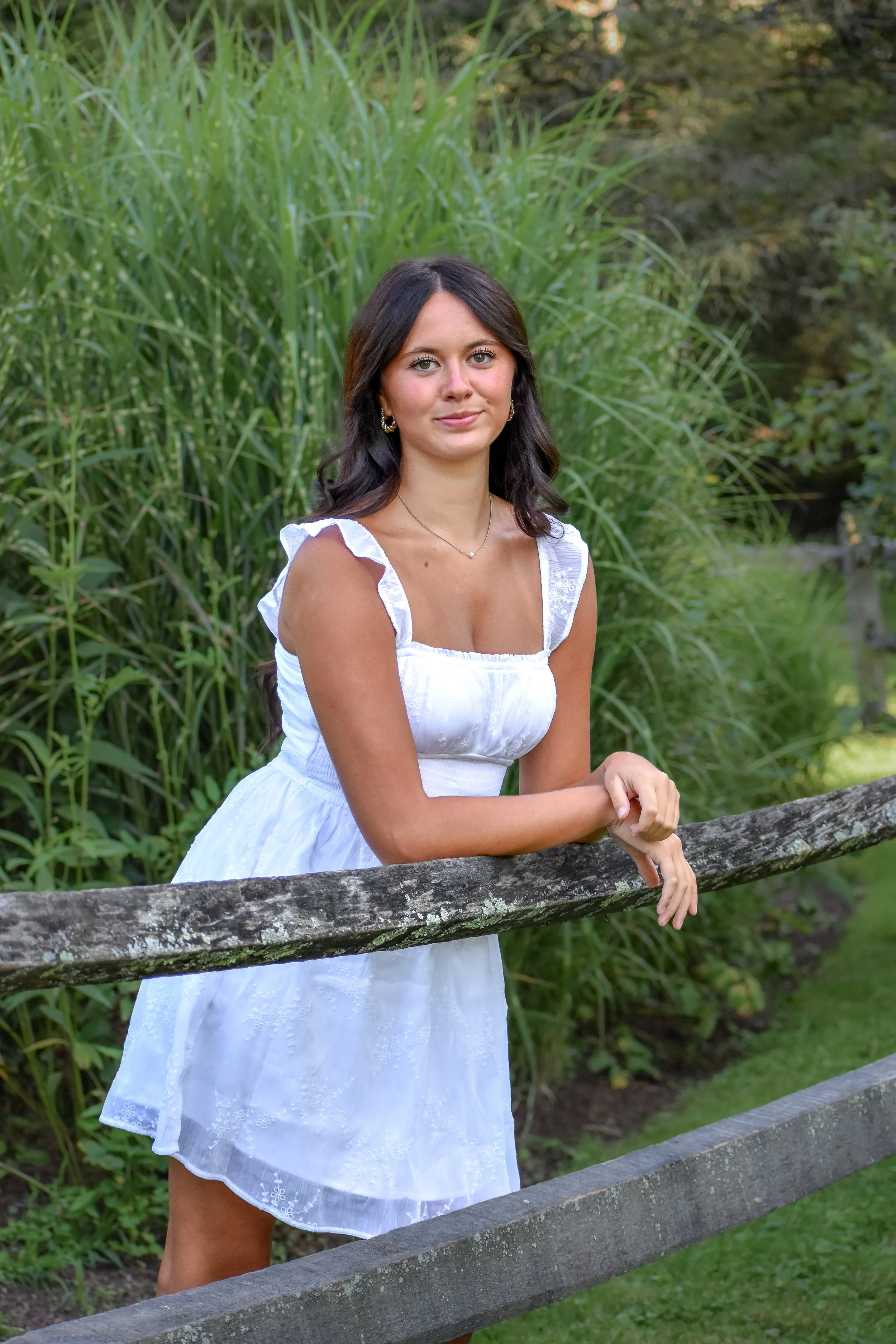 A young woman in a white dress standing outdoors near a wooden fence, with green grass and tall plants in the background.