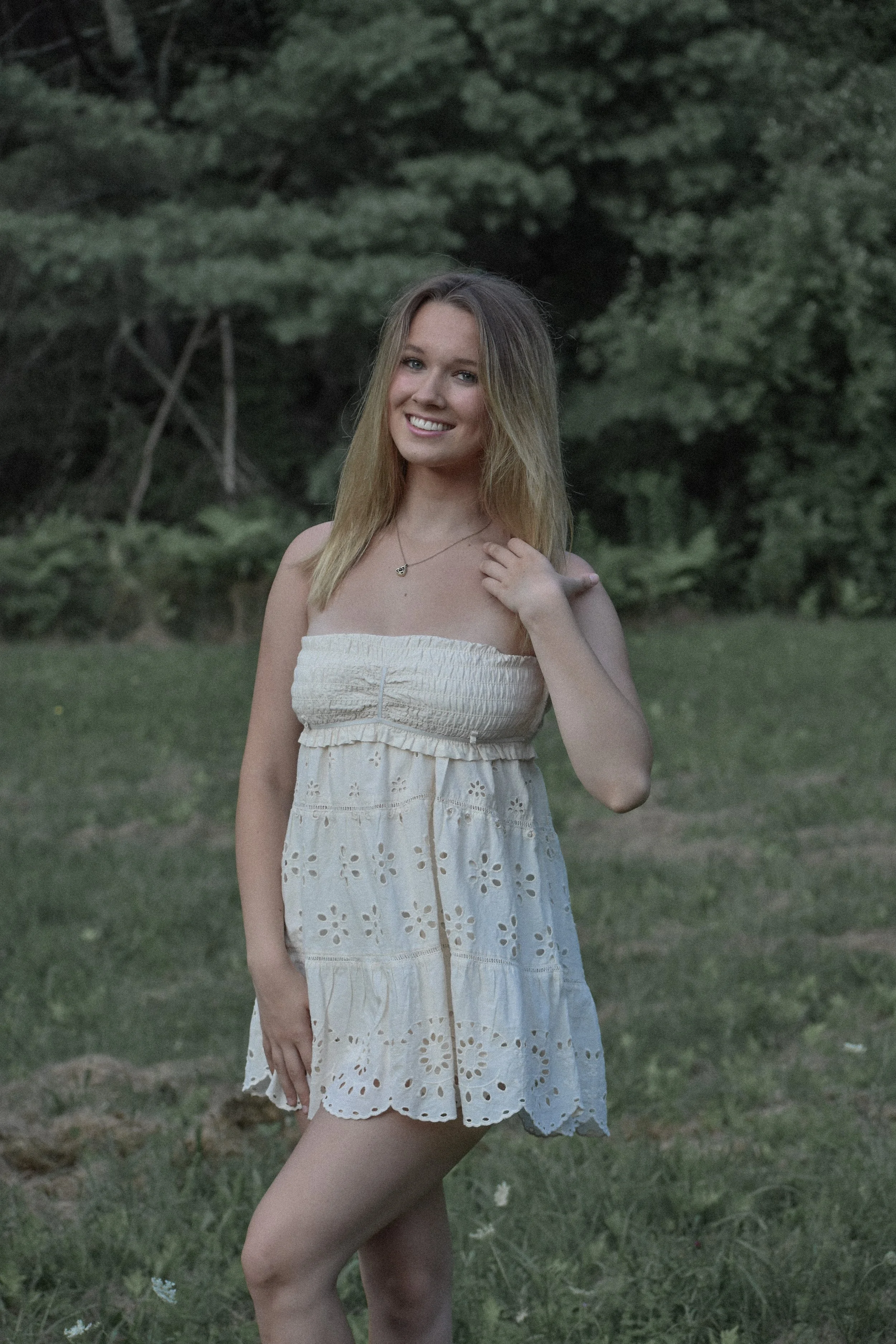 A young woman with blonde hair in a strapless white dress standing outdoors in a grassy area with trees in the background, smiling.