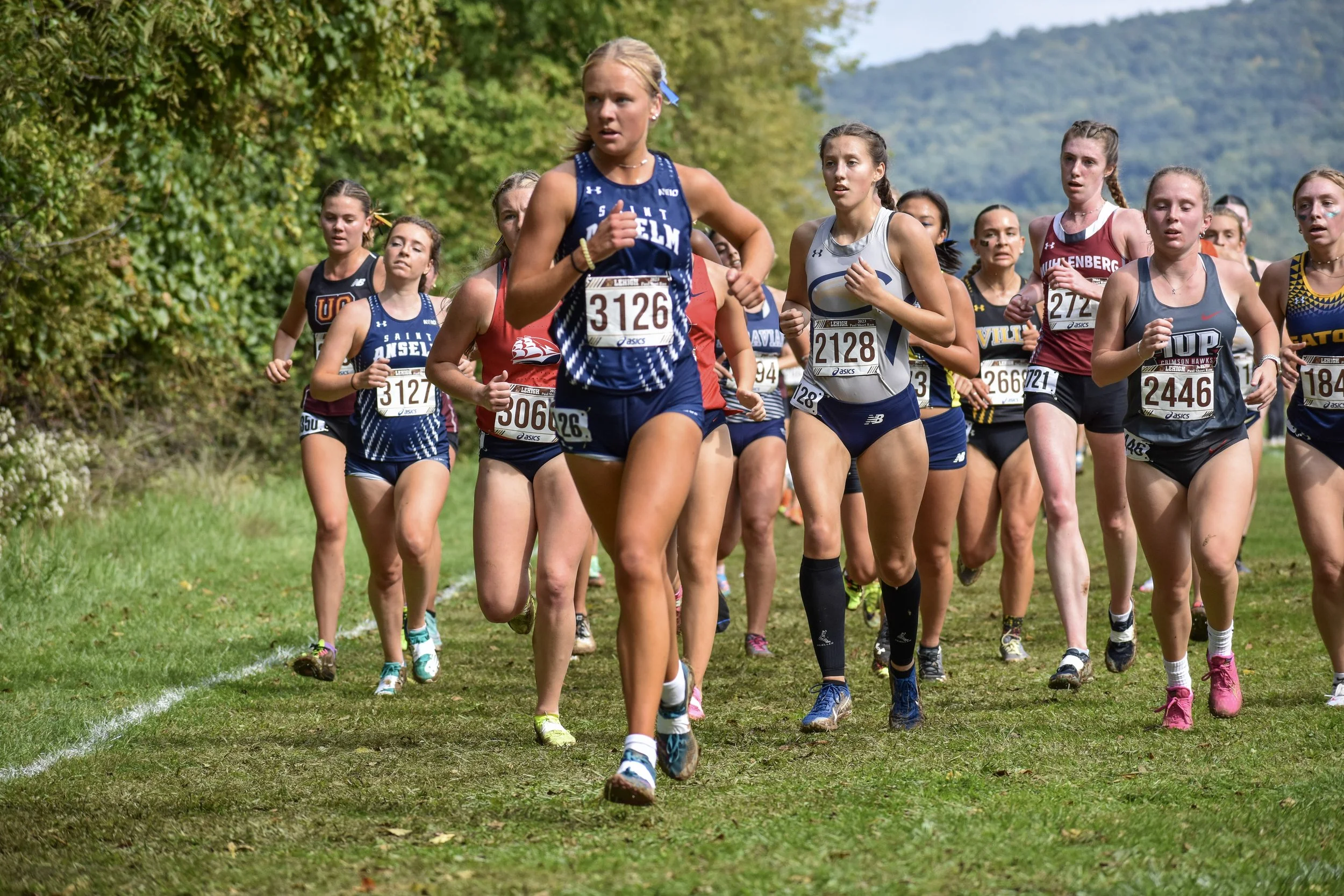 Group of female cross country runners competing in a race on a grassy trail surrounded by trees with hills in the background.