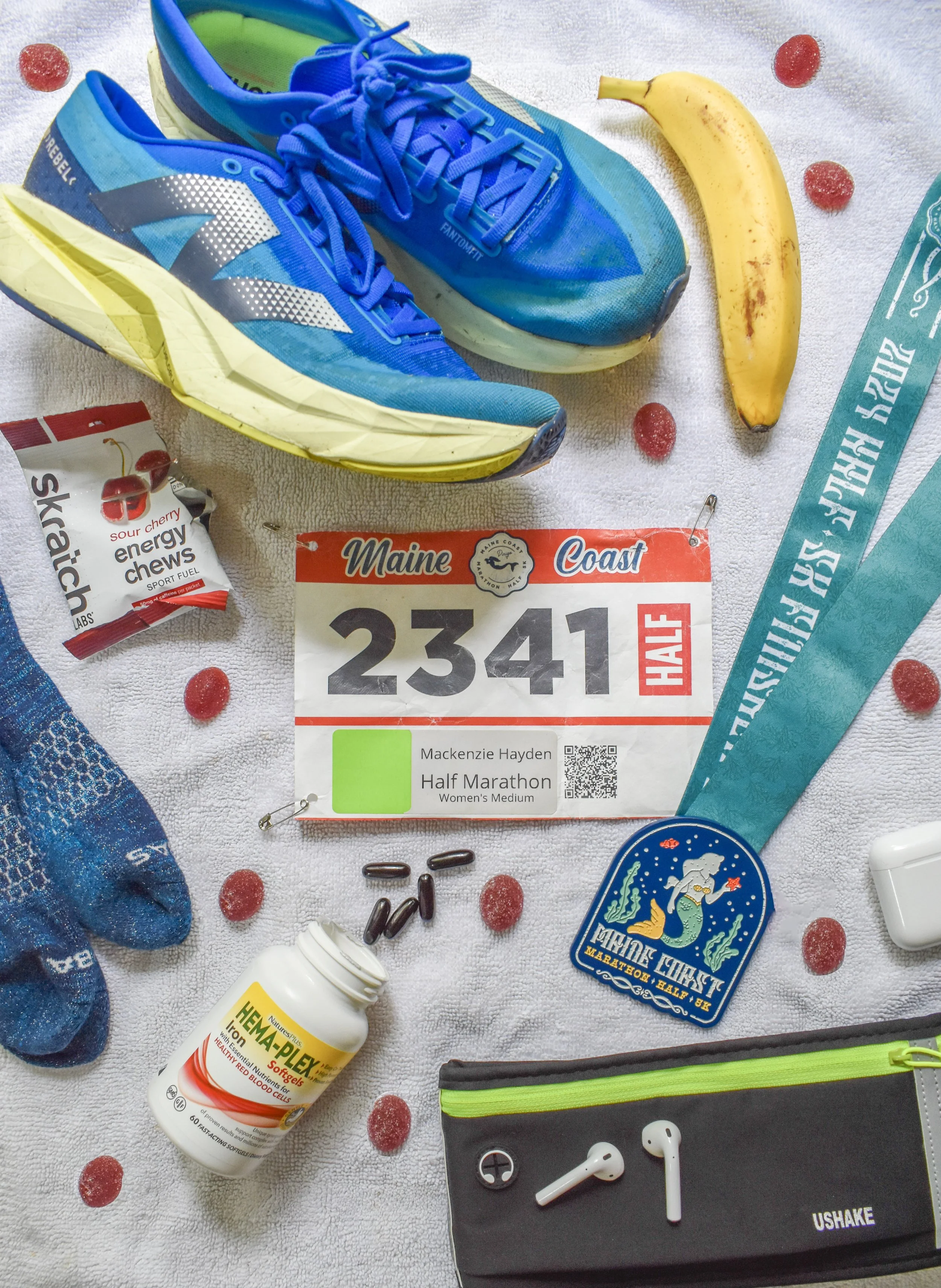 Items for a half marathon, including blue running shoes, a yellow banana, energy chews, a race bib with number 2341, a blue laminated map, a patch, vitamin pills, earbuds in a case, and Gatorade containers on a white towel with gummy candies.