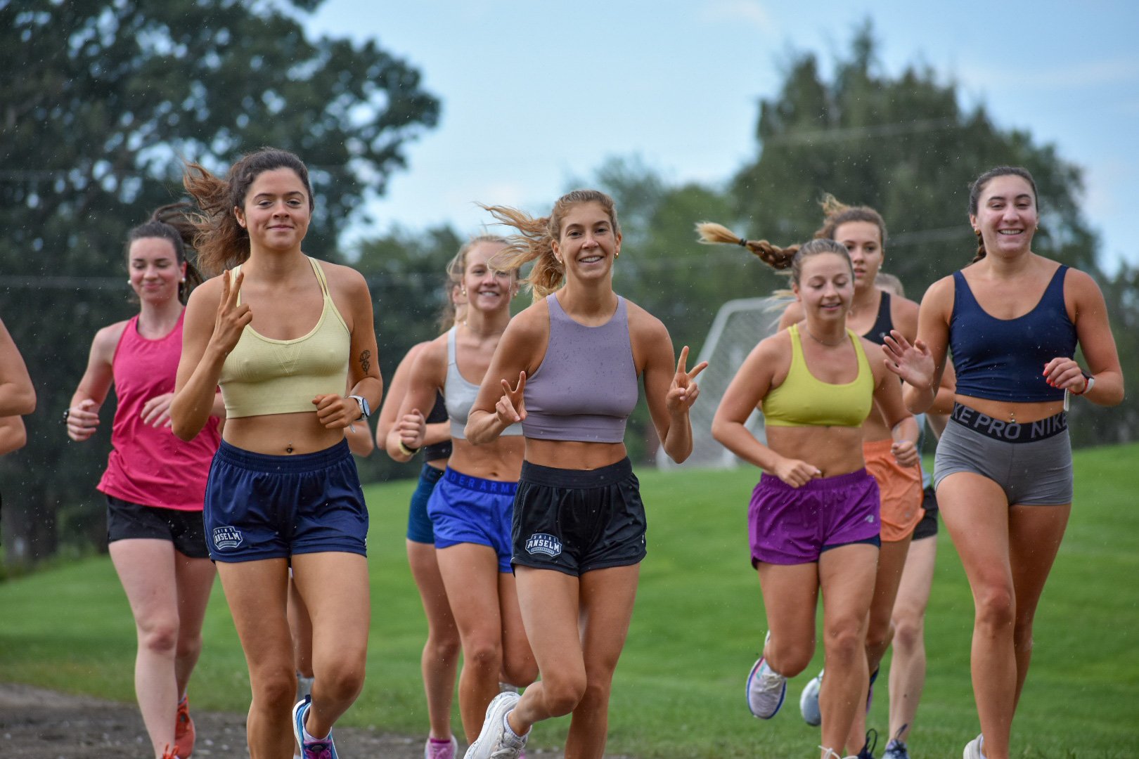 Group of women running outdoors on a grassy field, some smiling and making peace signs, wearing athletic clothing.
