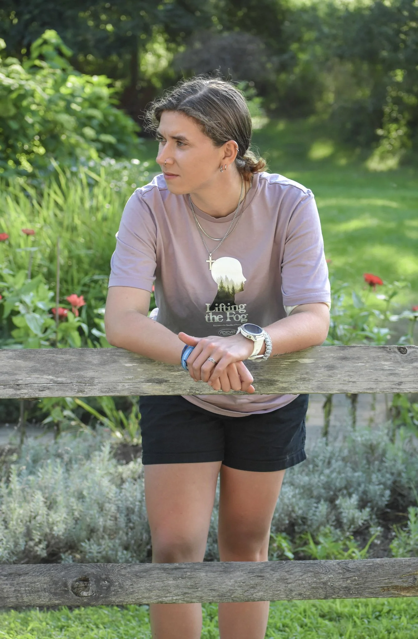 A woman leaning on a wooden fence outdoors, looking to the side with a thoughtful expression, surrounded by greenery and plants.