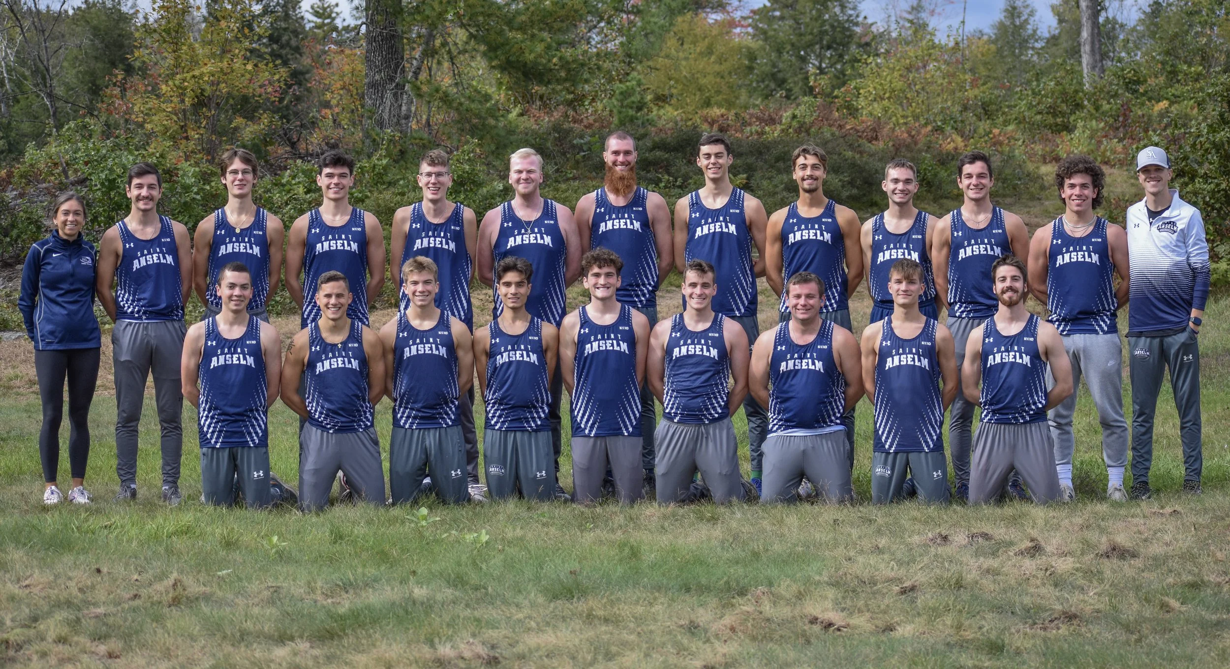 A group of young male and female track athletes with two coaches posed outdoors on grass, standing in front of trees with autumn foliage. They are wearing blue and white uniforms with 'Saint Anselm' on the front.
