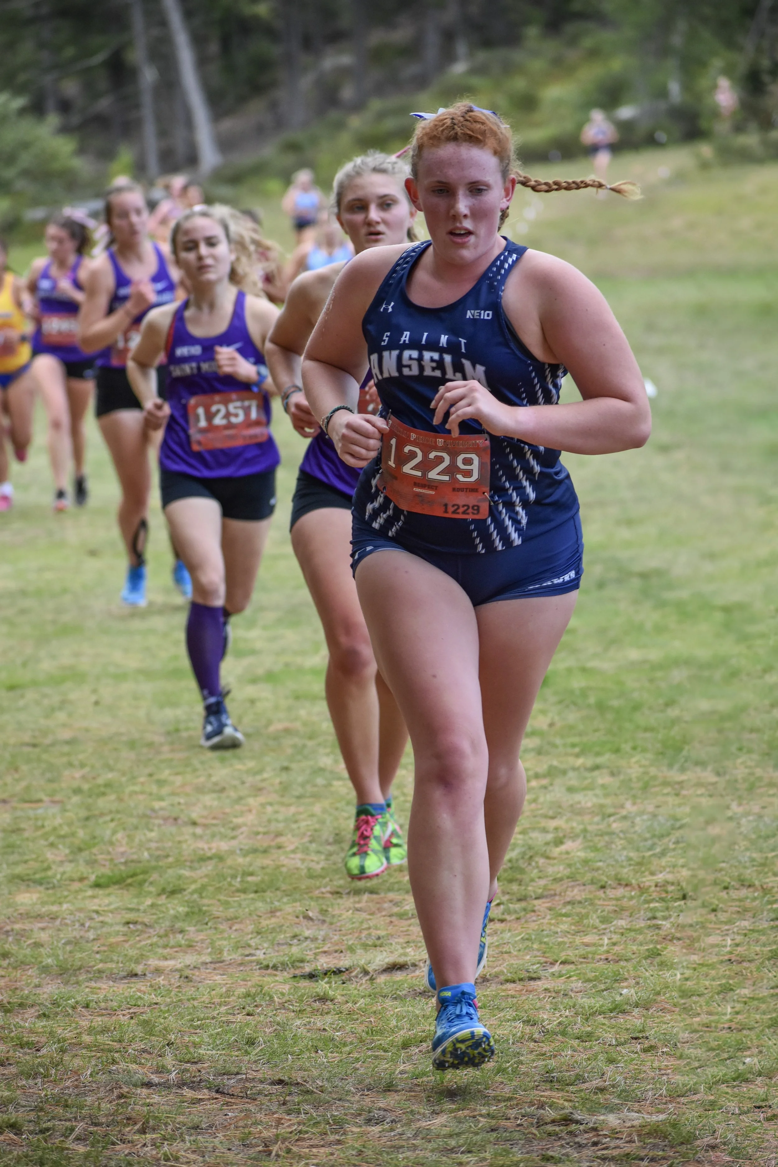 A group of female runners competing in a cross-country race outdoors on grass with trees in the background.