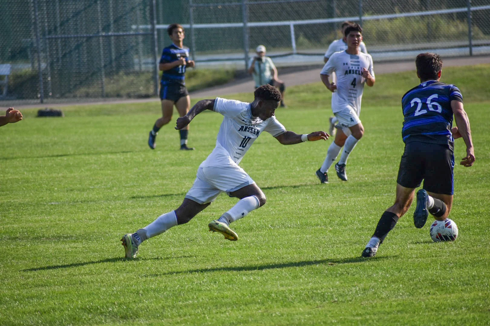 Soccer players in action during a match on a sunny day on a green grass field, with a chain-link fence and some spectators in the background.