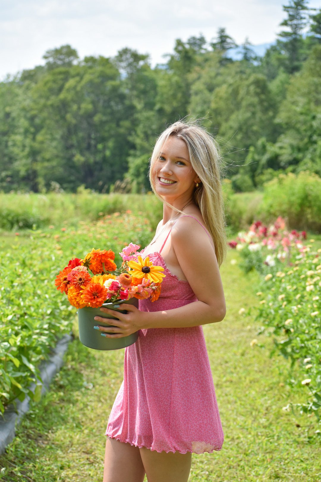 A young woman with long blonde hair wearing a pink dress holds a pot of colorful flowers and stands in a flower field with green trees in the background.