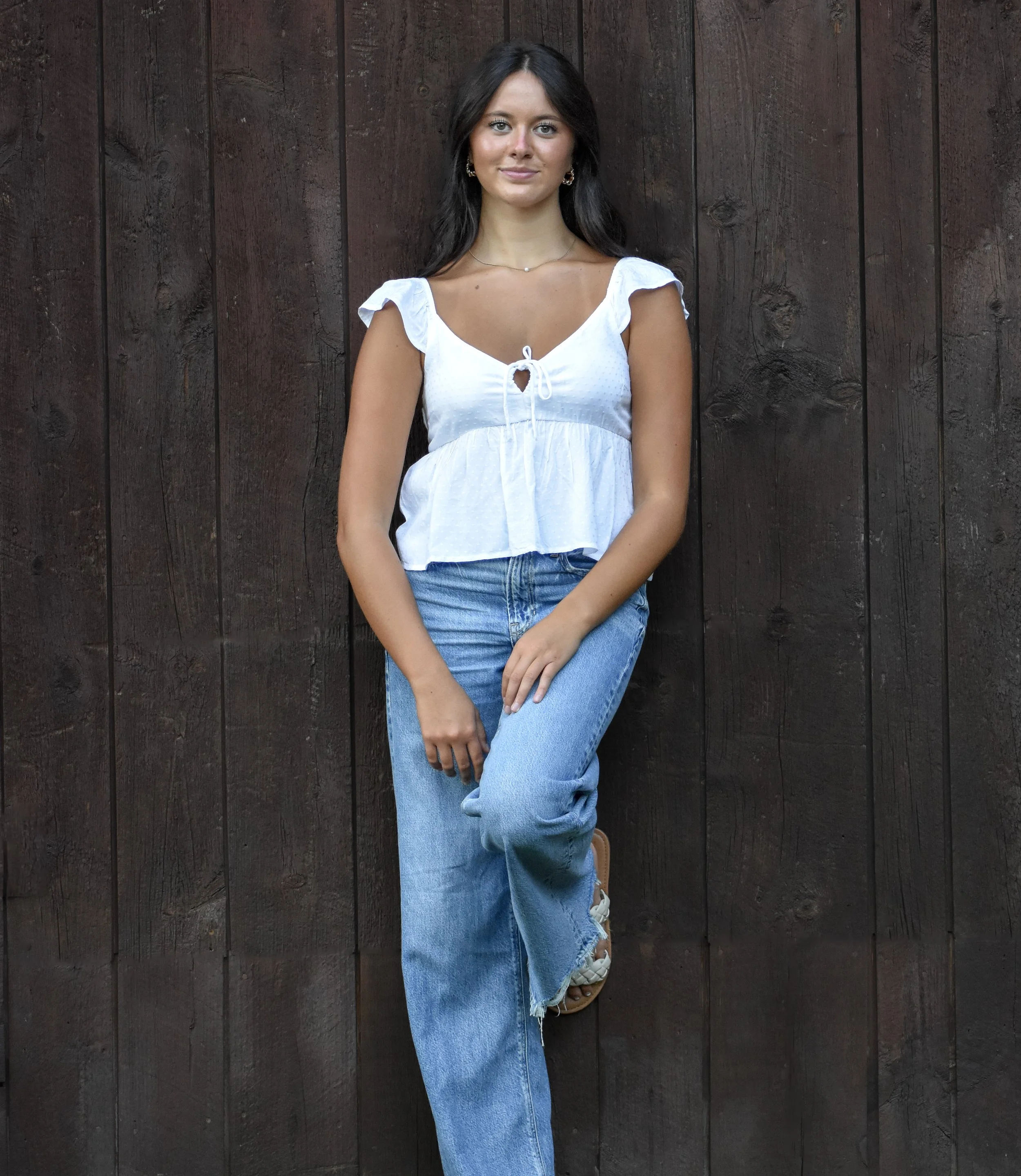 A woman standing against a dark wooden wall, wearing a white ruffled top, blue jeans, and sandals.