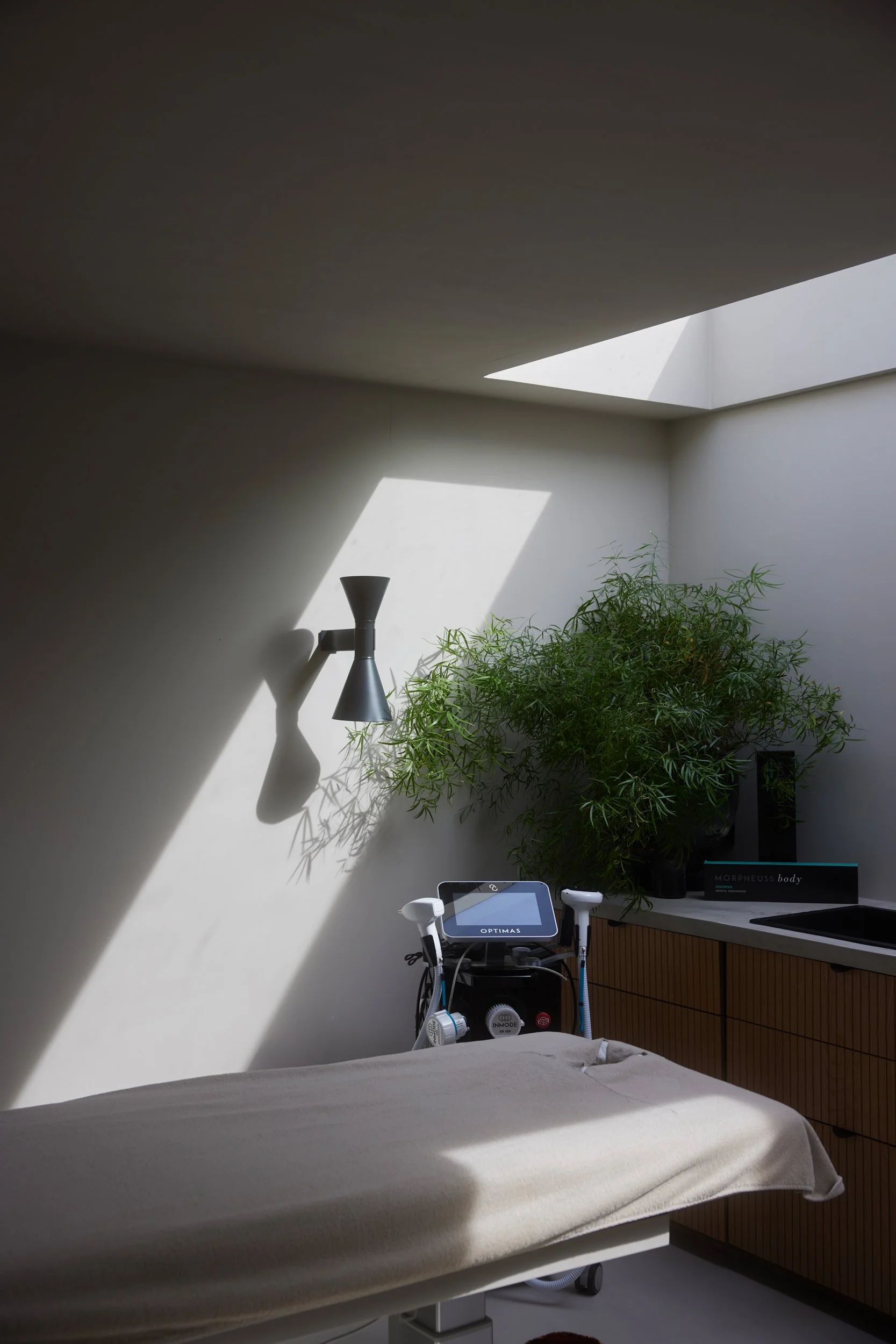 Een kalme behandelkamer met een futon, medische apparatuur, planten en een grote raam met natuurlijk licht.