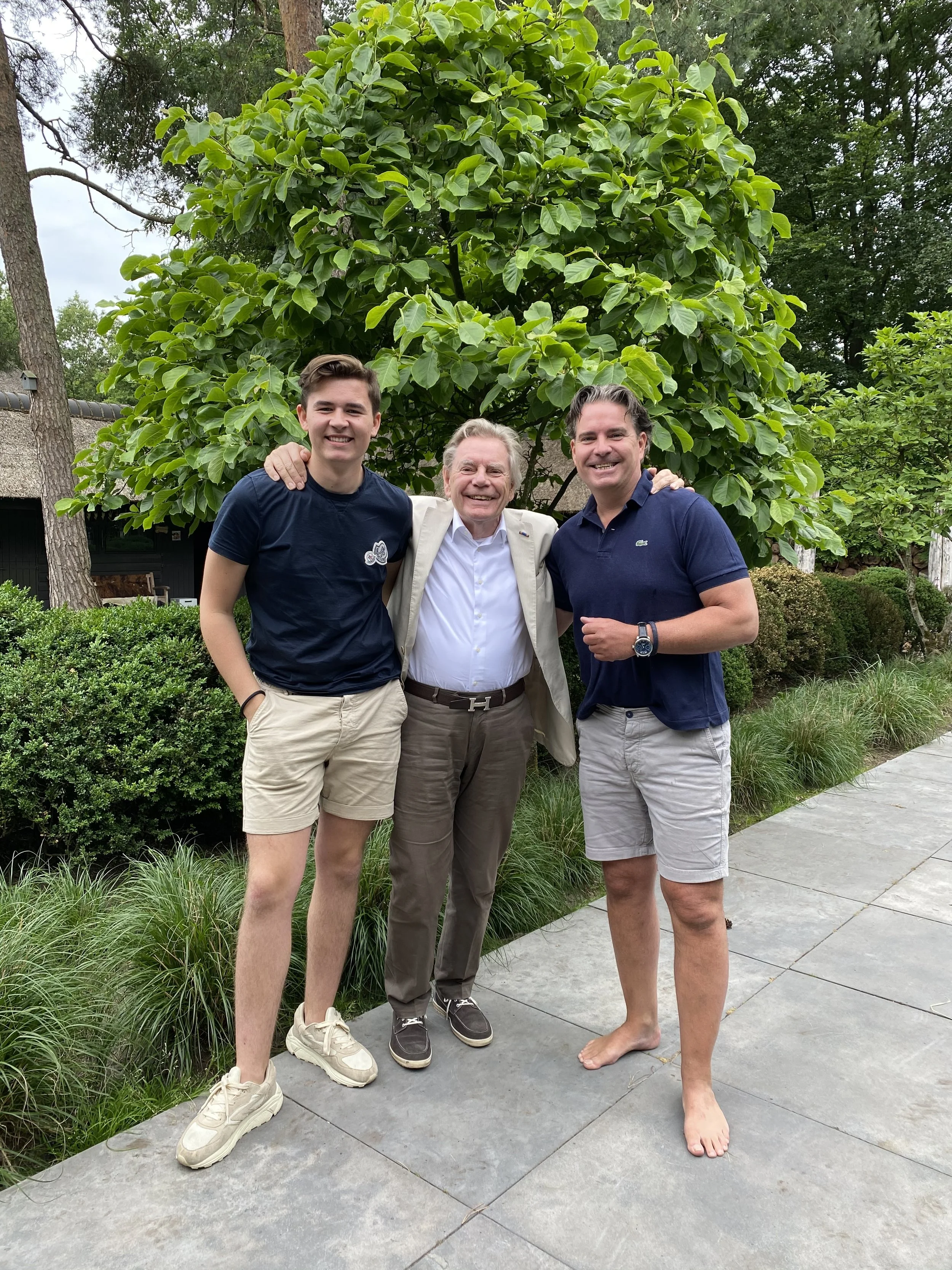 Drie mannen staan samen buiten voor een groene boom, lachend en met armen om elkaar. Twee dragen korte broek en sneaker, de man in het midden draagt een beige jas en bruin broek.