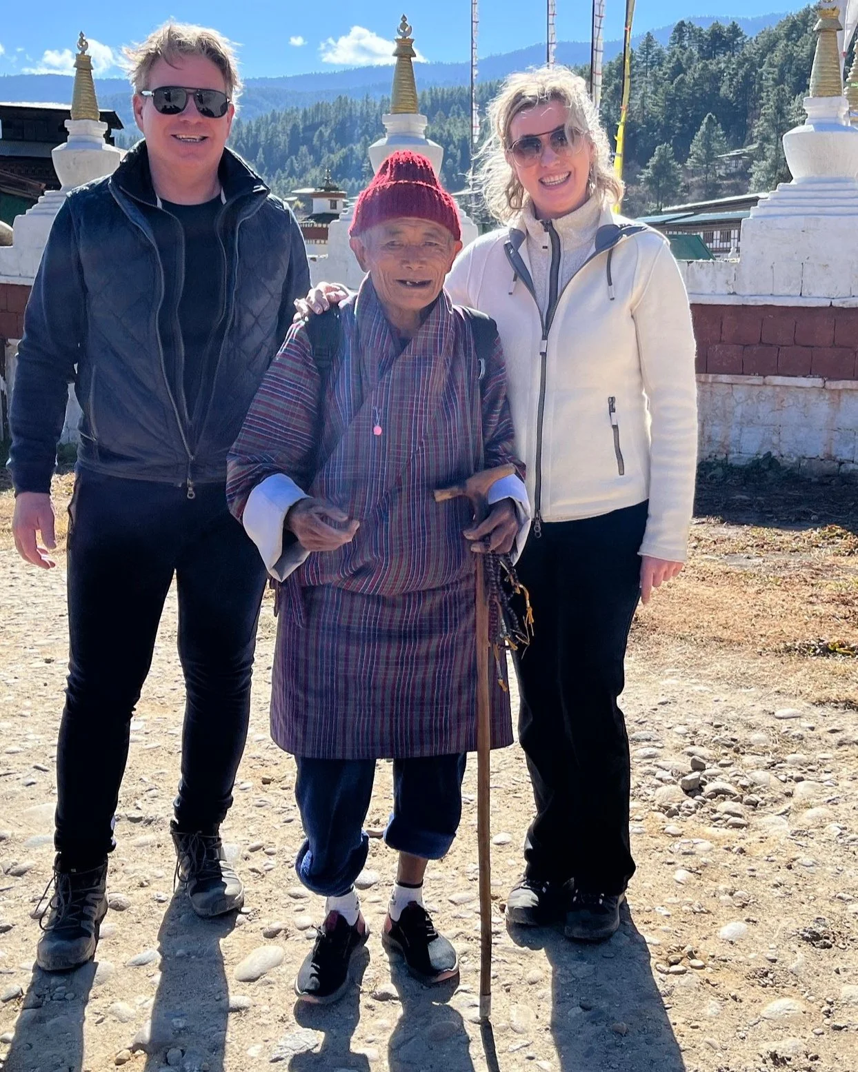Twee jonge toeristen, een man en een vrouw, poseert samen met een oudere lokale man, die een traditionele kleding draagt en een wandelstok vasthoudt, voor een tempel in een bergachtig gebied.