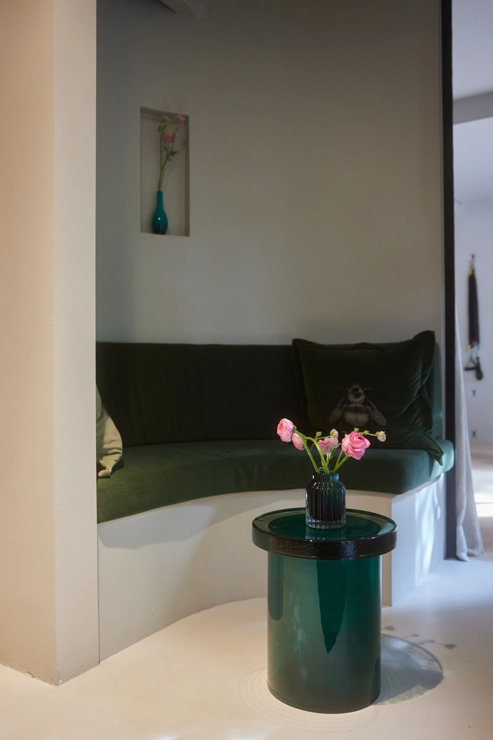 Binneninterieur met een groen bank, een ronde groene tafel met een vaas met roze bloemen en een gesneden muur met nis met een blauwe vaas en bloemen.