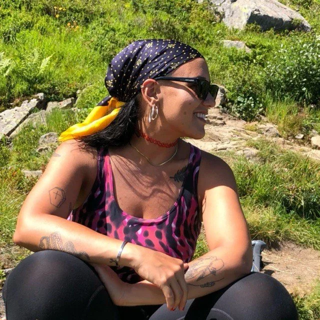 A woman sitting outdoors on a grassy hillside, smiling and looking to the right. She is wearing sunglasses, a black bandana with yellow polka dots, a pink and black animal print top, and various jewelry, with visible tattoos on her arms.