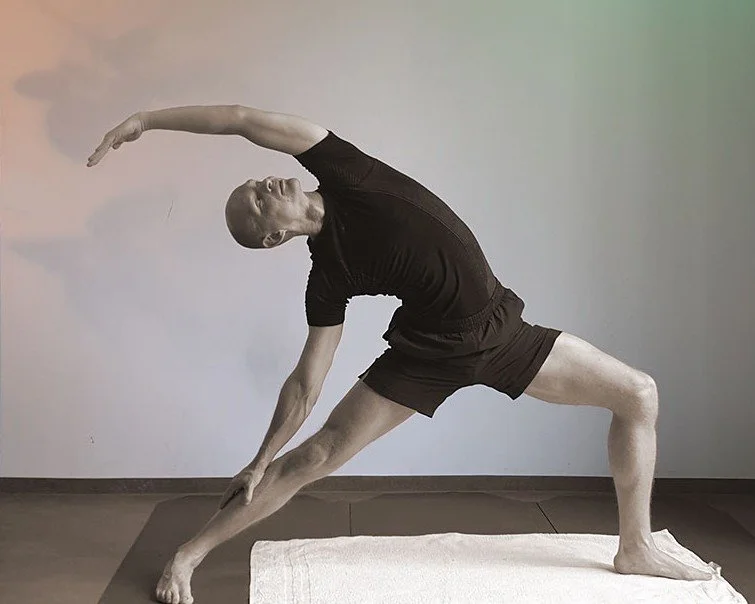 A man practicing yoga indoors, demonstrating a side angle stretch with one arm extending over his head and the other hand touching the floor.