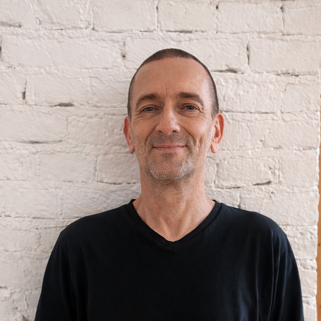 A man with a shaved head and light skin wearing a gray sweater, standing against a plain gray background.