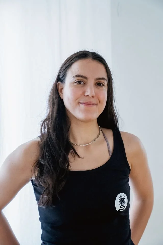 A woman with long brown hair wearing a black sleeveless top with a white logo, standing in front of a plain white background.