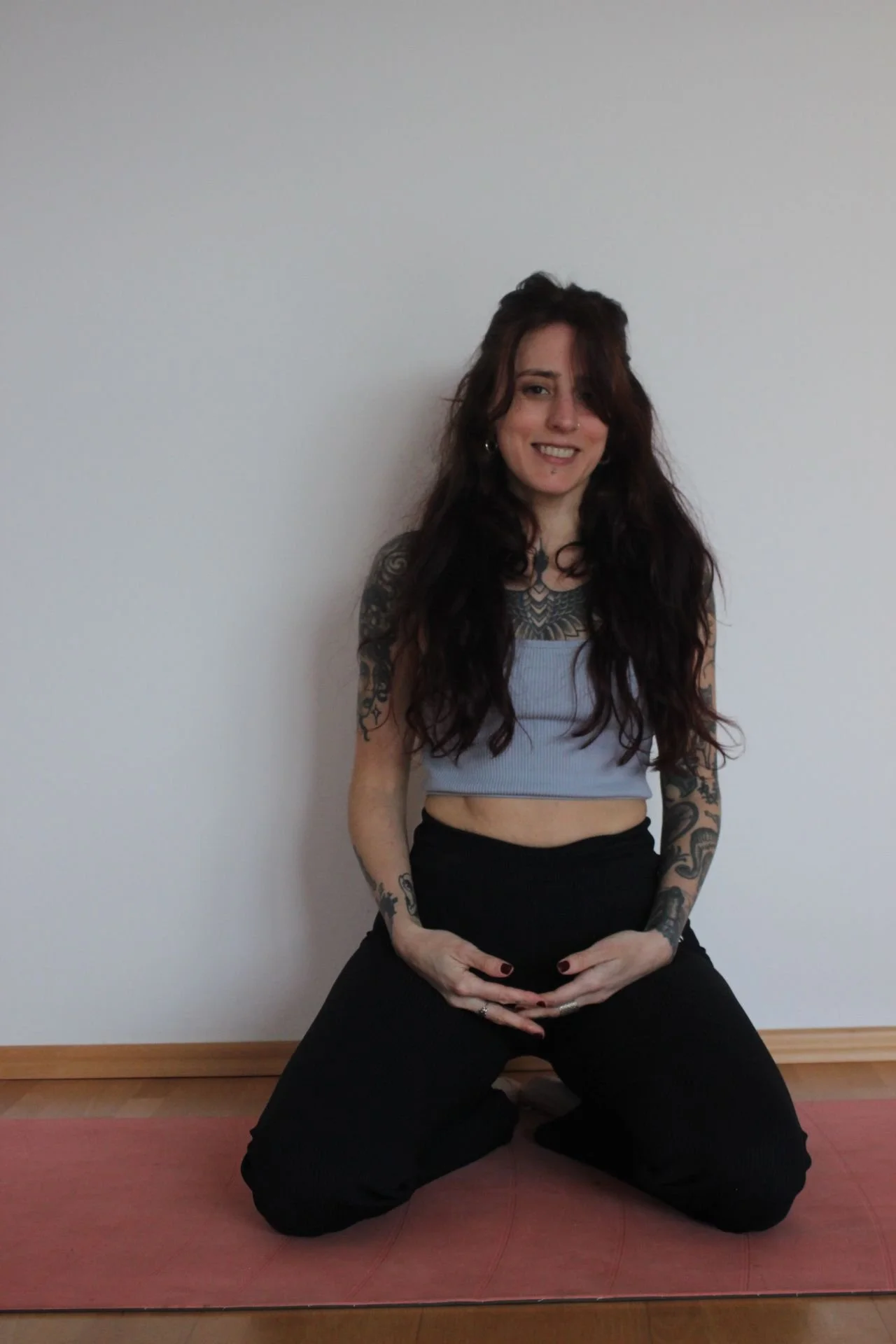 A woman with long dark wavy hair and tattoos on her arms, sitting on a yoga mat on the floor, smiling at the camera. She is wearing a light blue crop top and black pants, with a white wall in the background.