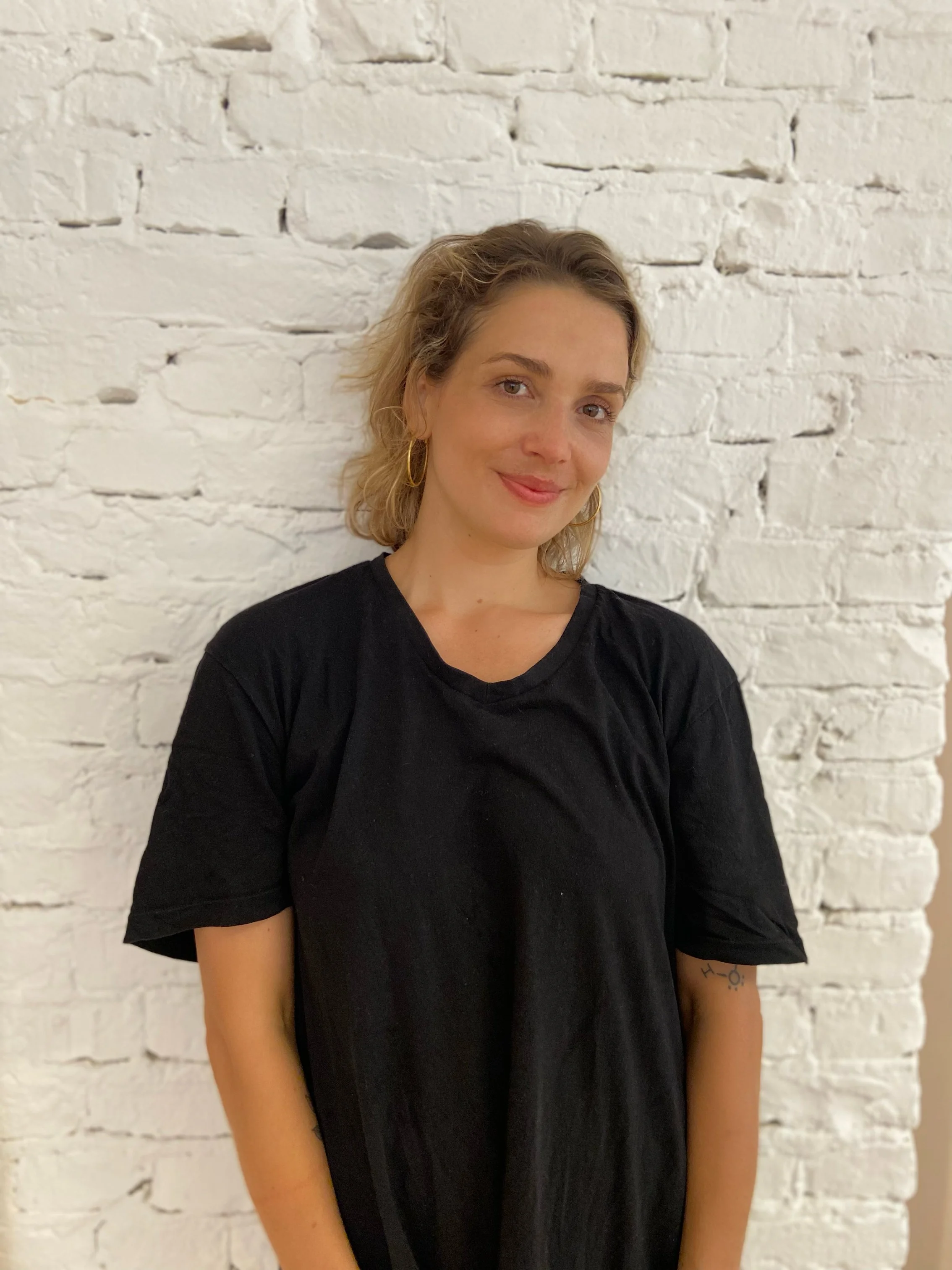 A woman with blonde tousled hair and gold hoop earrings standing in front of a white brick wall, smiling slightly, wearing a loose black t-shirt.