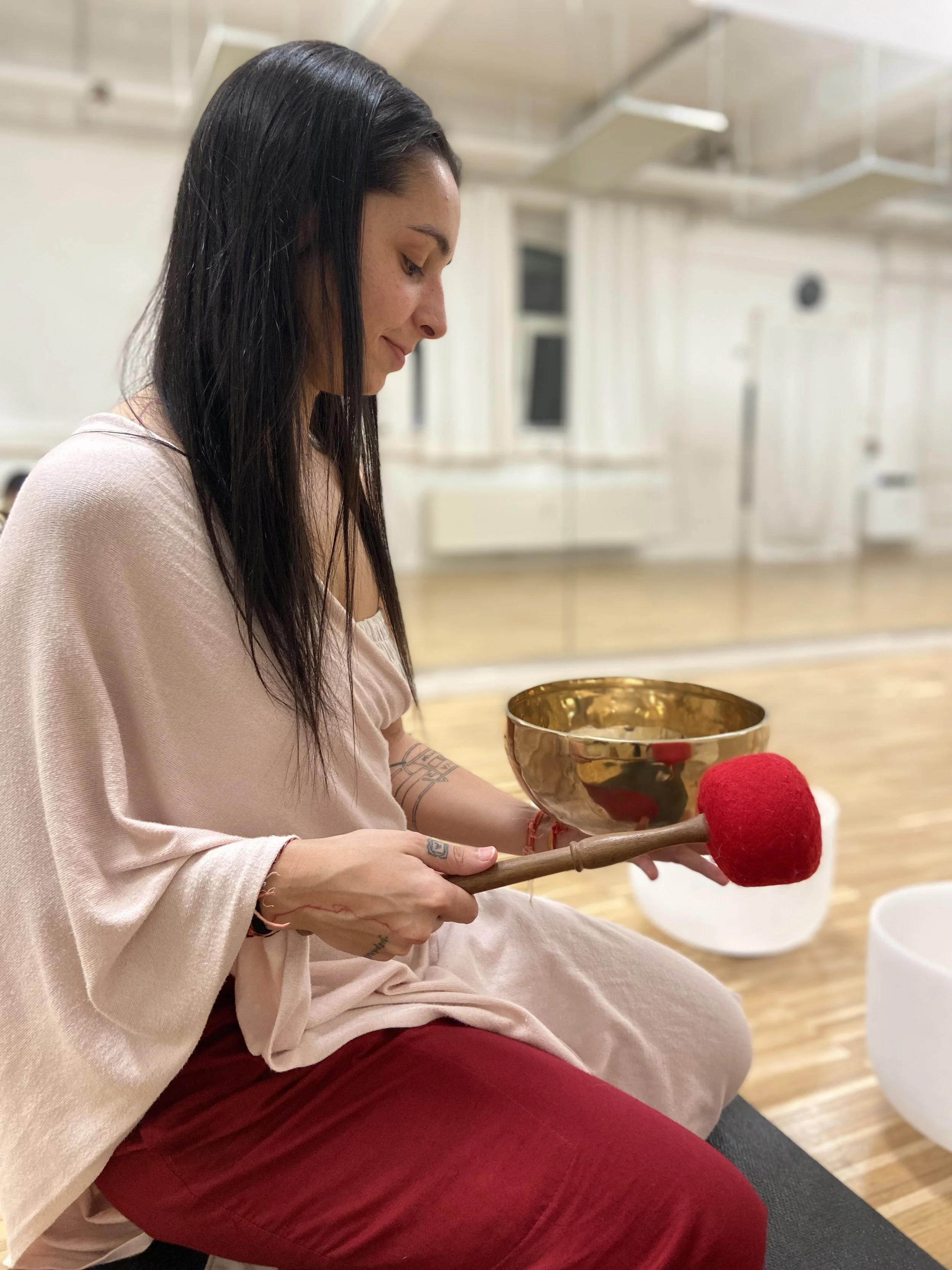 A woman with long dark hair playing a singing bowl with a mallet in a room with wood floors and mirrors.