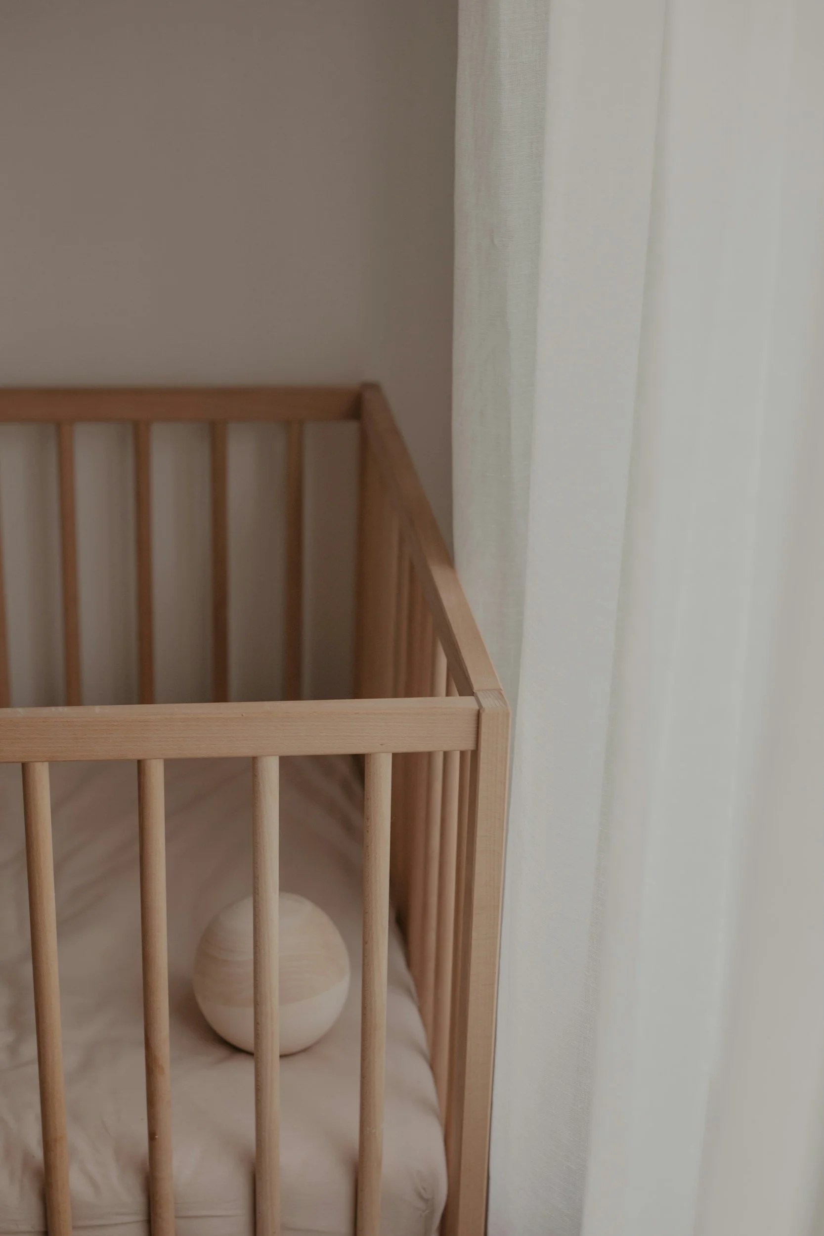 A wooden baby crib with vertical slats, lined with white fabric and a white ball inside, next to a sheer white curtain.