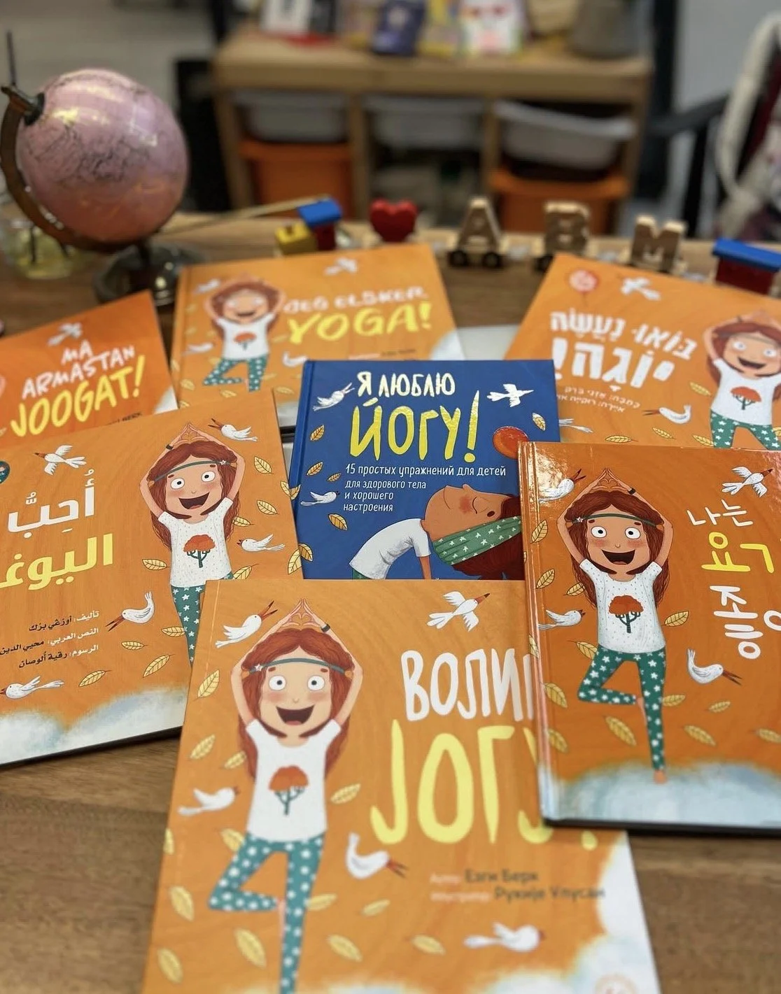 Renkli çocuk yogası kitaplarıyla dolu masa, üzerinde çeşitli şekillerde yoga yapan çocukların çizimleri bulunuyor. Kitapların önemli bölümü turuncu renkte olmakla birlikte mavi ve diğer renkler de mevcut. Ayrıca masa üzerinde küçük dekoratif nesneler