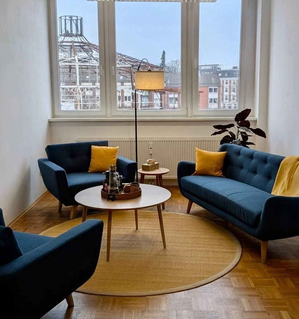 Gemütliches Wohnzimmer mit drei blauen Sofas, gelben Kissen, einem runden Teppich, einem kleinen Beistelltisch, einem Stehlampe und einer großen Fensterfront mit Blick auf ein Stadtbild.