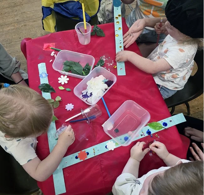 Children engaging in a craft activity at a table covered with a red tablecloth, using various art supplies like paper, flowers, and decorative stickers. Making may flower head bands.