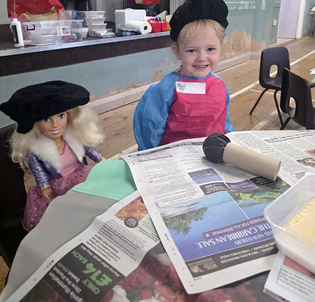 A young girl wearing a black chef's hat and pink apron, smiling at a table with a doll, craft supplies, in a room with a counter and chairs.