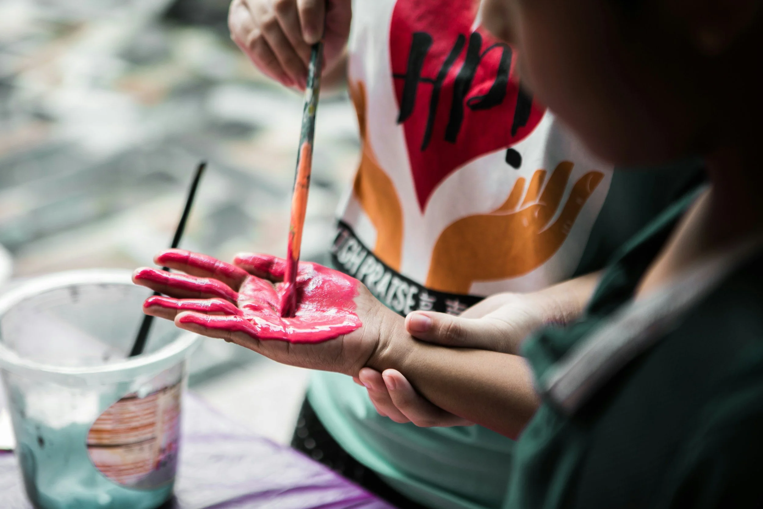 A person is holding a pink-colored ice cream cone in their hand, and they are painting on their hand with a paintbrush. The person is wearing a shirt with a red, black, white, and orange design. Several cups of ice cream are visible in the background.