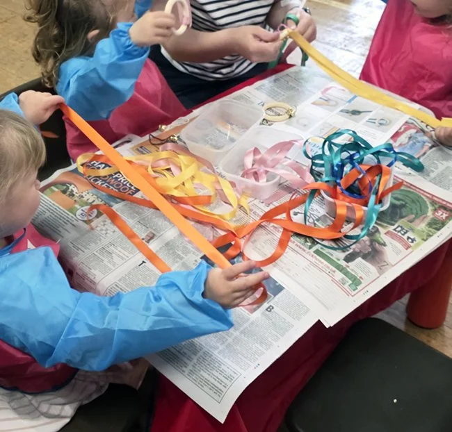 Children at a craft table working with colorful ribbon strips and scissors, on the table.
