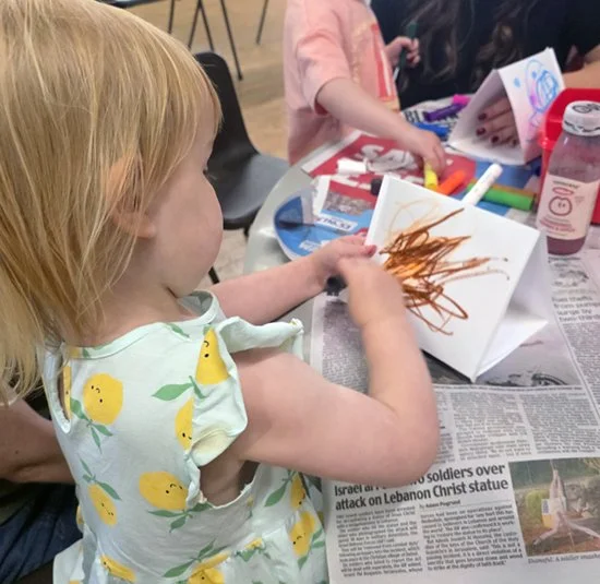 A young girl with red hair drawing with brown marker on a blank paper folded like a tent, at a table with other children, surrounded by art supplies.