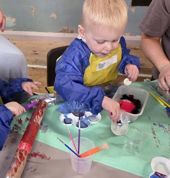 A young boy with blond hair and a blue and yellow smock is engaging in a painting activity at a table, surrounded by art supplies, including paintbrushes, a plastic container with paints, and a cardboard tube covered in red paint.