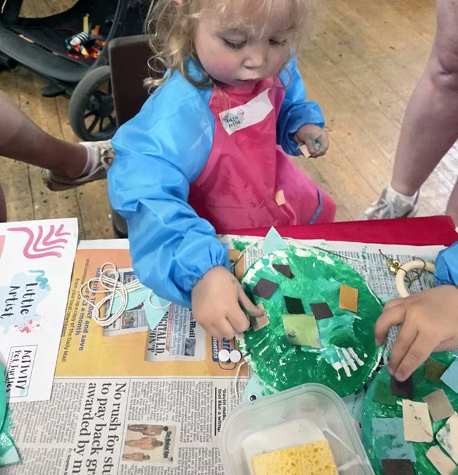 A young girl in a blue shirt and pink aprons working on a craft project at a table, decorated a green paper turtle with colourful paper squares, with craft supplies nearby.