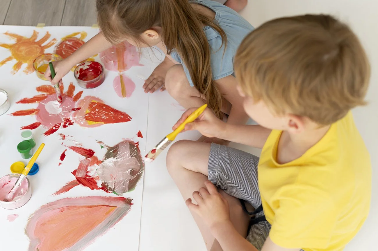 Two children painting colorful abstract art on a large sheet of paper with brushes and paint.